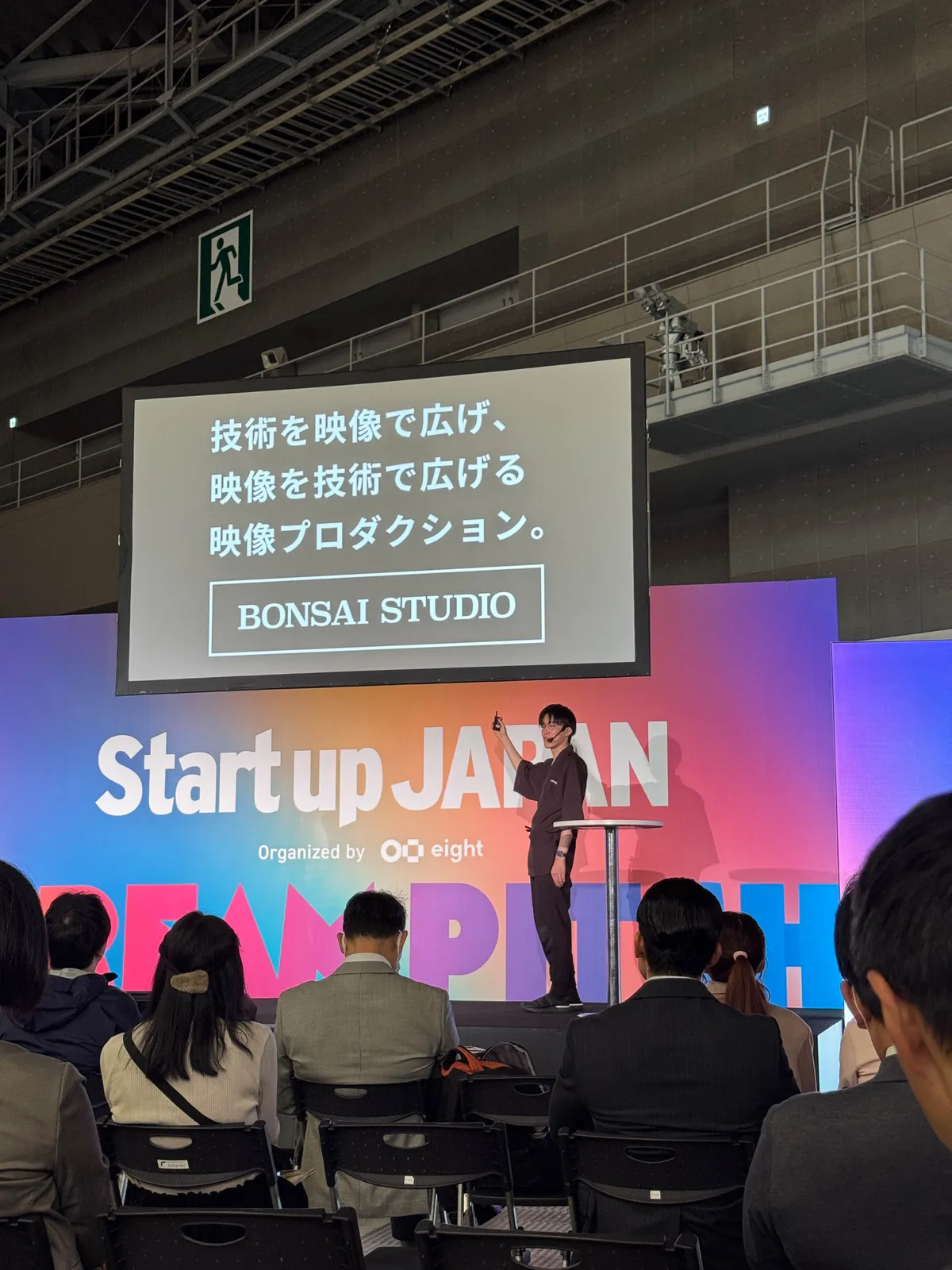 ステージ上に大きく「Start up JAPAN」と表示されたスクリーンがあり、その前で黒い服を着た男性がプレゼンテーションを行っている。スクリーンには「BONSAI STUDIO」の文字も映し出されている。