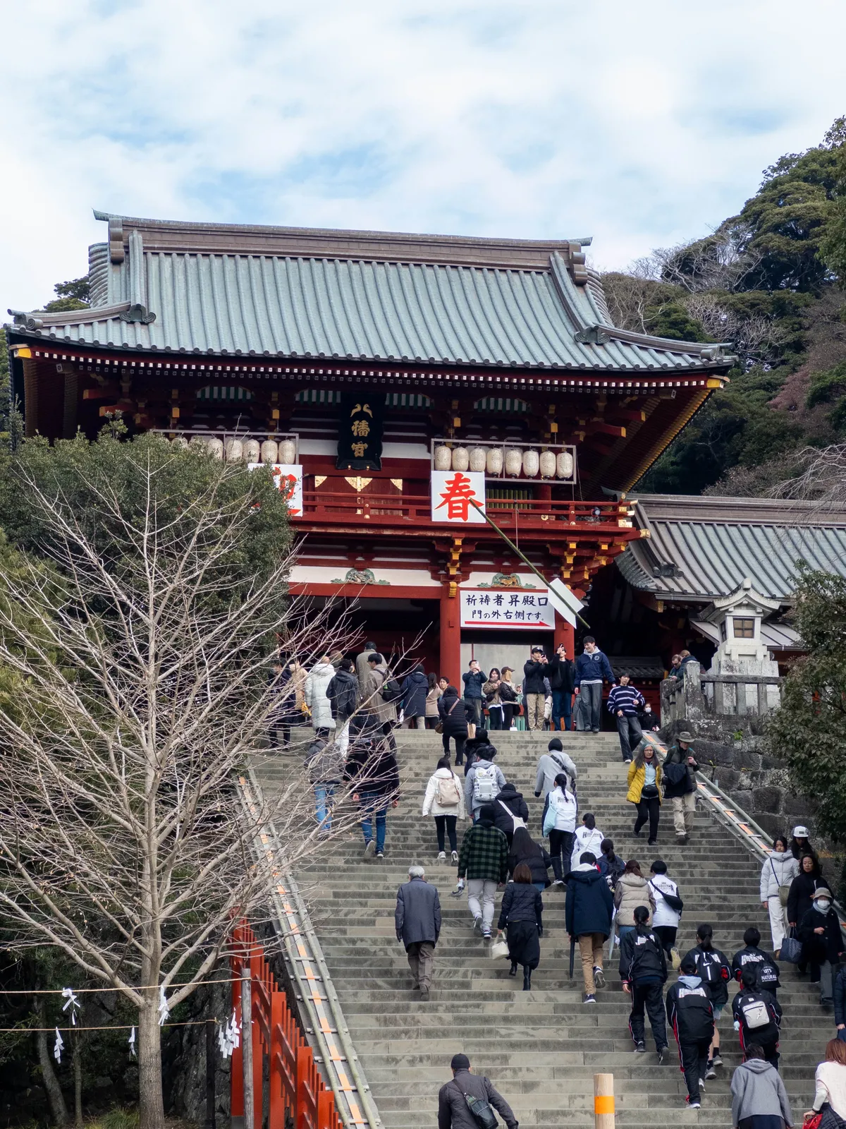 朱色の大きな神社の門に続く長い石段を、多くの参拝客が登っている。社殿の前には「春」などの文字が飾られ、初詣や参拝の様子がうかがえる。