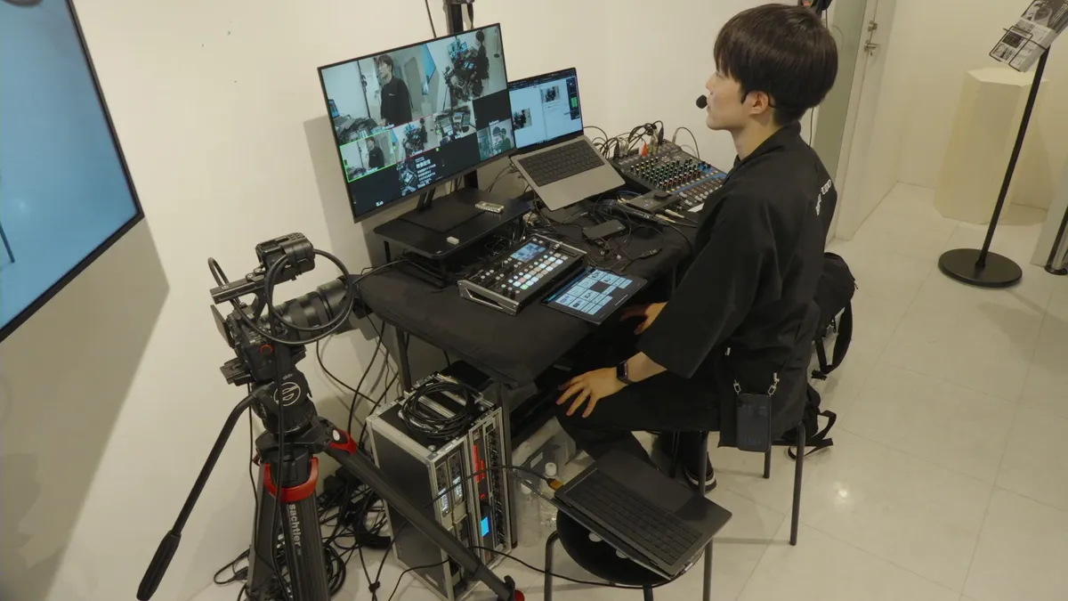 配信卓の前でライブ配信について解説する鈴木一平。モニター、カメラ、音声機器が並んでいる。