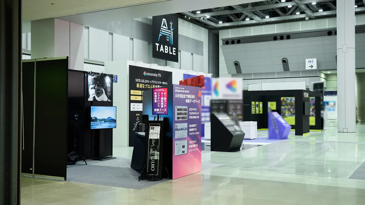 展示会場の通路からの風景。天井に「AI TABLE」のバナーが吊るされ、手前には「CINEMATICA」の赤いパネルのブース、奥には他社のブースが並んでいる。