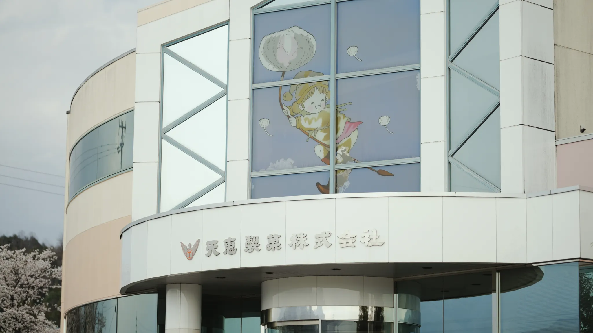 建物のエントランス部分。窓にはキャラクターのイラストがあり、建物には「天恵製菓株式会社」の文字が見える。