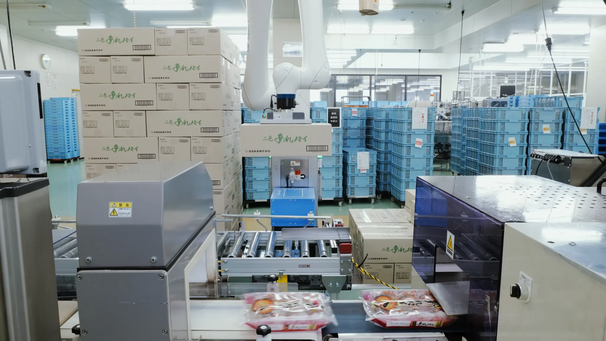 食品工場内で稼働するロボットアームとコンベアシステム。手前では個包装された製品が流れ、奥には段ボール箱や青いカゴが多数積み上げられている。
