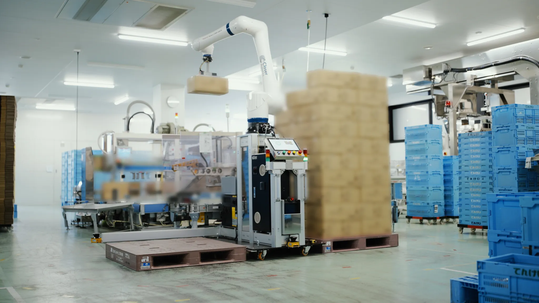 Closer Roboticsのロゴがある白いロボットアームが、工場内で段ボール箱をパレットに積んでいる。