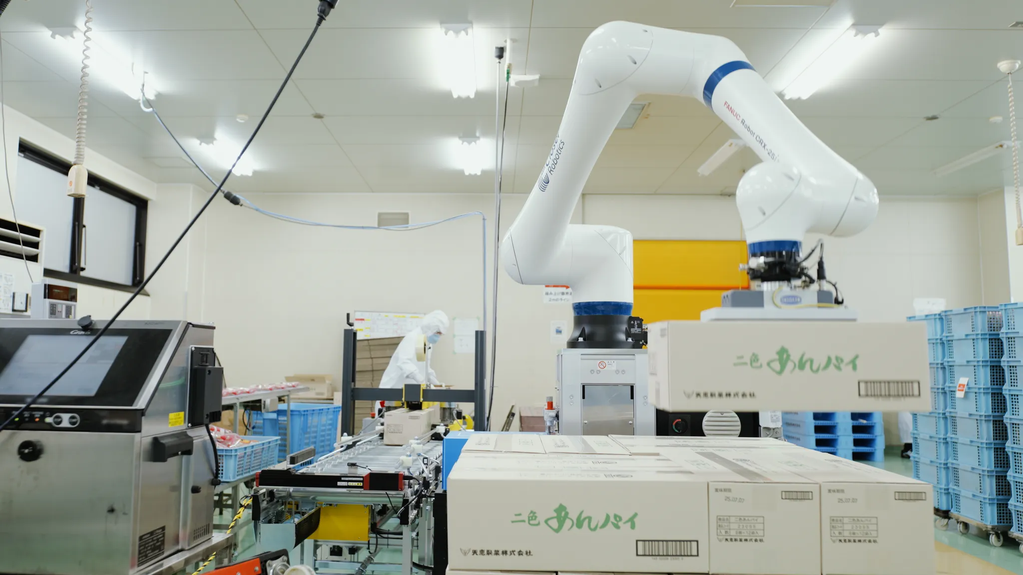 工場内で、FANUC Robot CRX-25iAと記されたCloser Roboticsの白いロボットアームが「二色あんパイ」と書かれた段ボール箱を運んでいる。奥には別の作業員と青いカゴが見える。