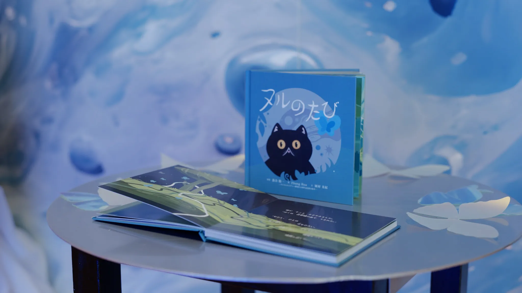 丸テーブルの上に、黒猫のイラストと日本語タイトル「ヌルのたび」が描かれた青い絵本が開かれた状態で展示されている。背景は青い抽象的な模様。