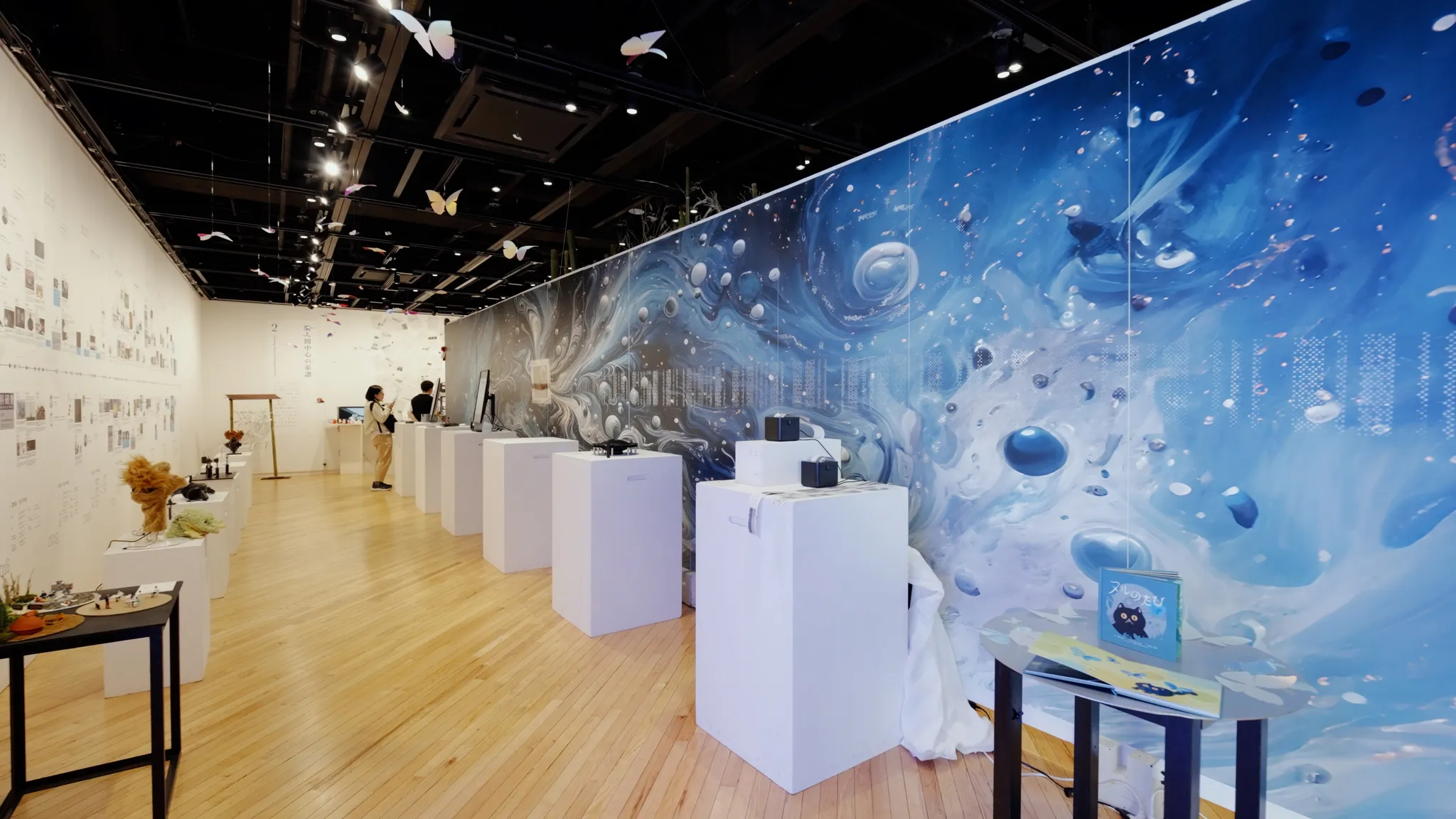 木目調の床に、白い展示台や様々な作品が並ぶ展示室。奥の壁には青異抽象的なグラフィックが大きく描かれ、天井からは蝶の飾りが多数ぶら下がっている。