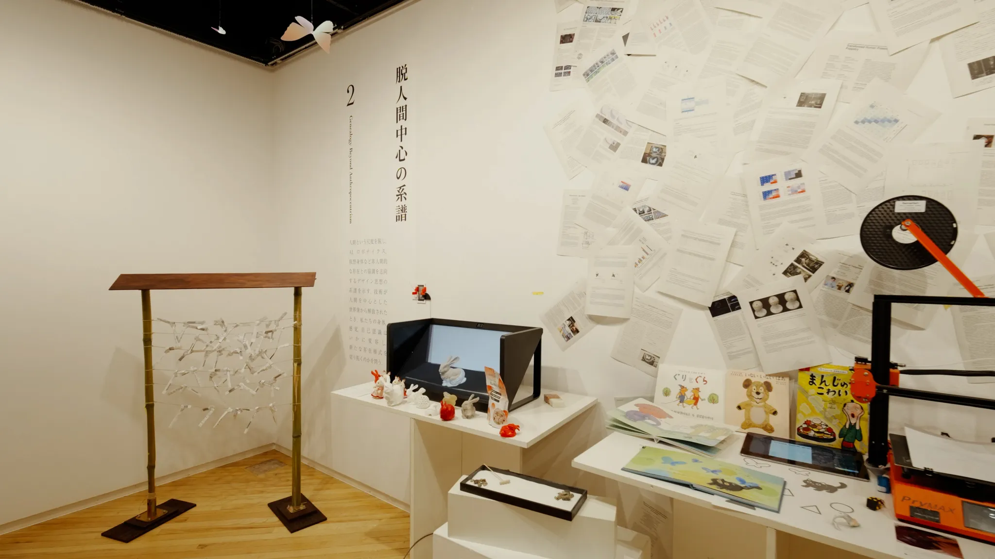 部屋の角にある展示。左には竹のフレームのおみくじ掛け、右には無数の論文が貼られた壁と、3Dプリンターなどが置かれた展示台がある。壁に文字「2 脱人間中心の系譜」