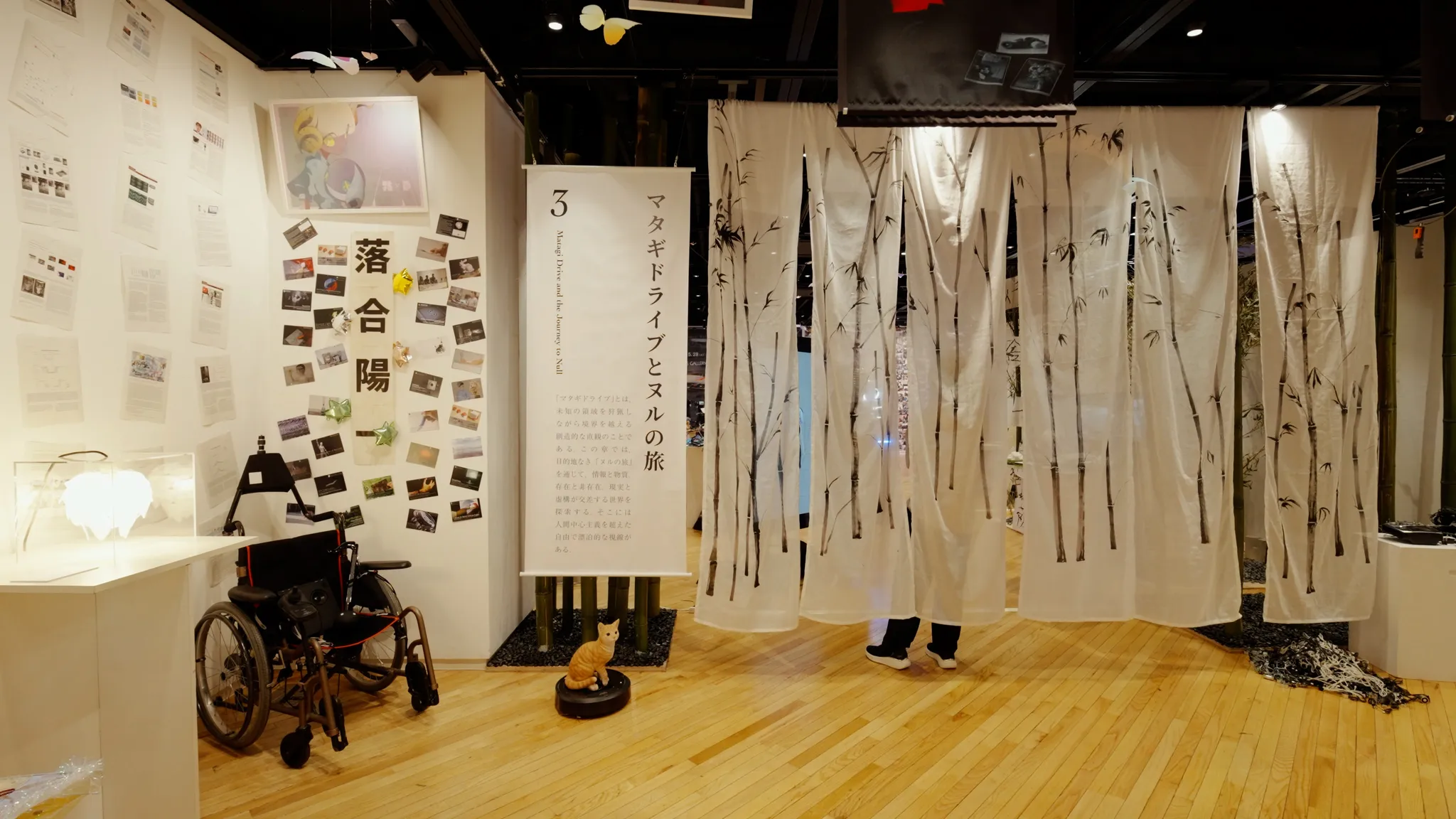 車椅子や、竹が描かれた白い布の暖簾が展示されたスペース。壁には多数の写真が貼られ、手前の床には猫の置物がある。パネル文字「3 マタギドライブとヌルお揃いの黒いロゴTシャツを着た2人の人物が立っている展示スペース。手前のデスクには様々な電子部品が並んでおり、奥の天井からは赤い傘などのオブジェがぶら下がっている。の旅」