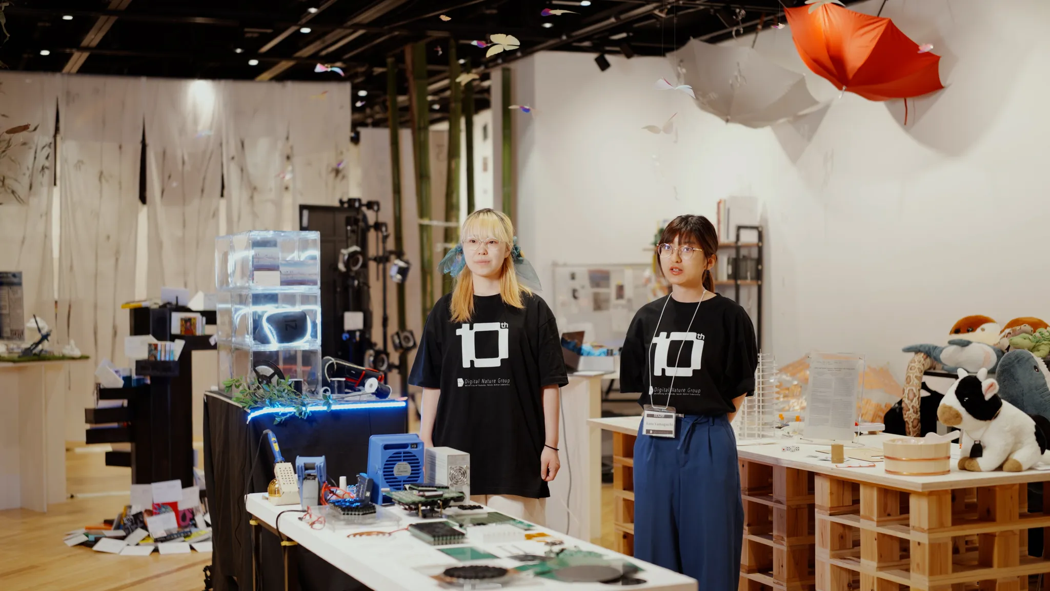 お揃いの黒いロゴTシャツを着た小澤知夏氏と山口果那氏が立っている展示スペース。手前のデスクには様々な電子部品が並んでおり、奥の天井からは赤い傘などのオブジェがぶら下がっている。