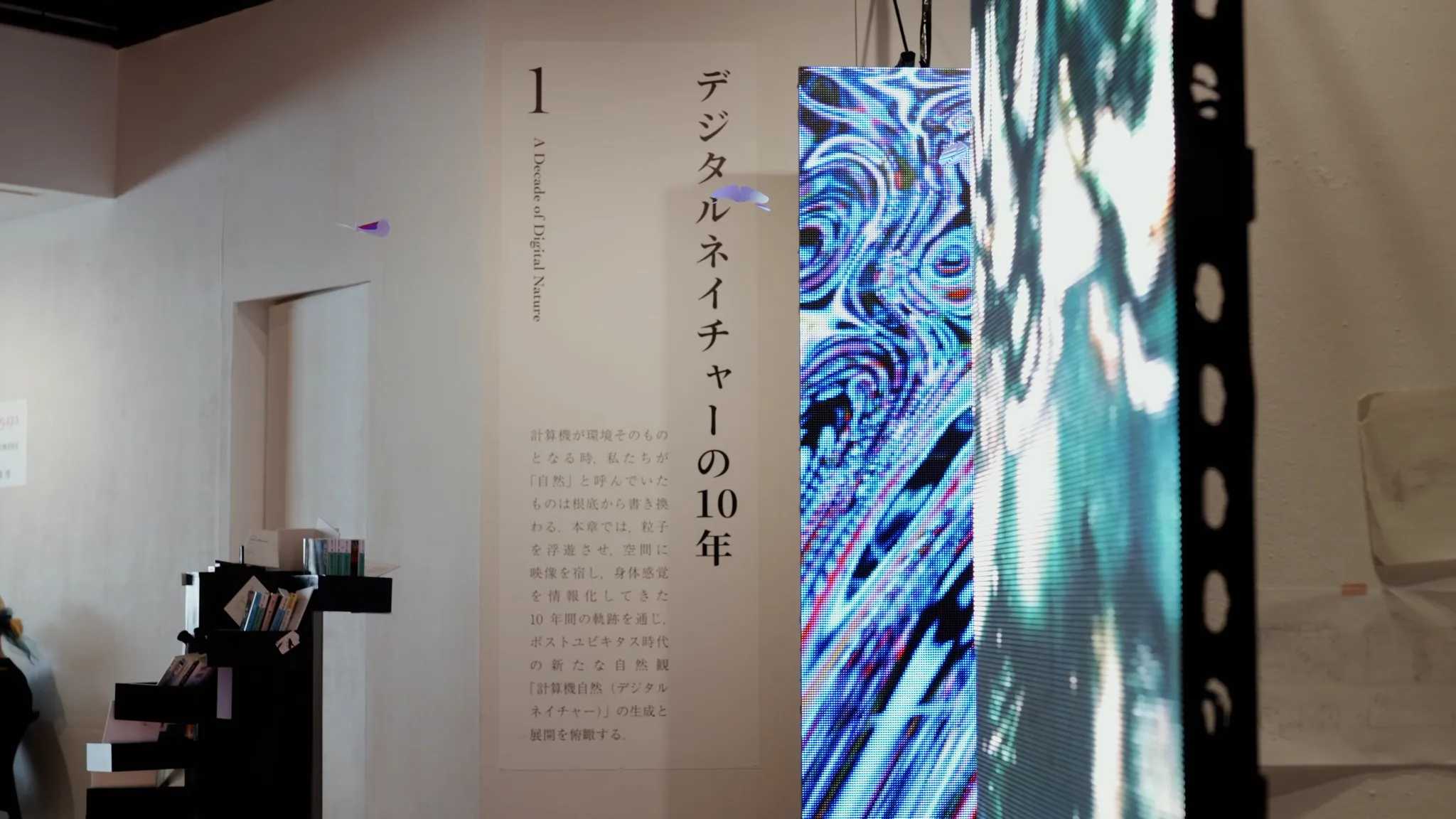 鮮やかな模様を映し出す縦型のデジタルスクリーンと、「1 デジタルネイチャーの10年 A Decade of Digital Nature」という英語と日本語のテキストが書かれた白い壁。左手には本が並べられた黒いスタンドがある。