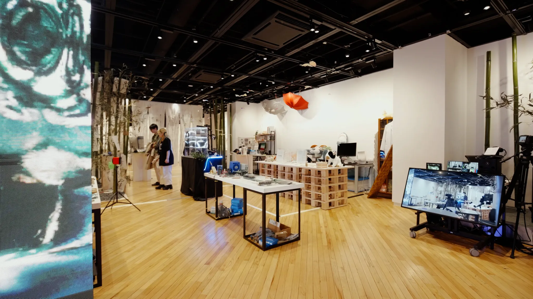 木目調の床が続く広い展示室の全体風景。手前にはLEDディスプレイのような柱やモニター付きの機材カートがあり、奥の展示スペースには見学している人物の姿が見える。