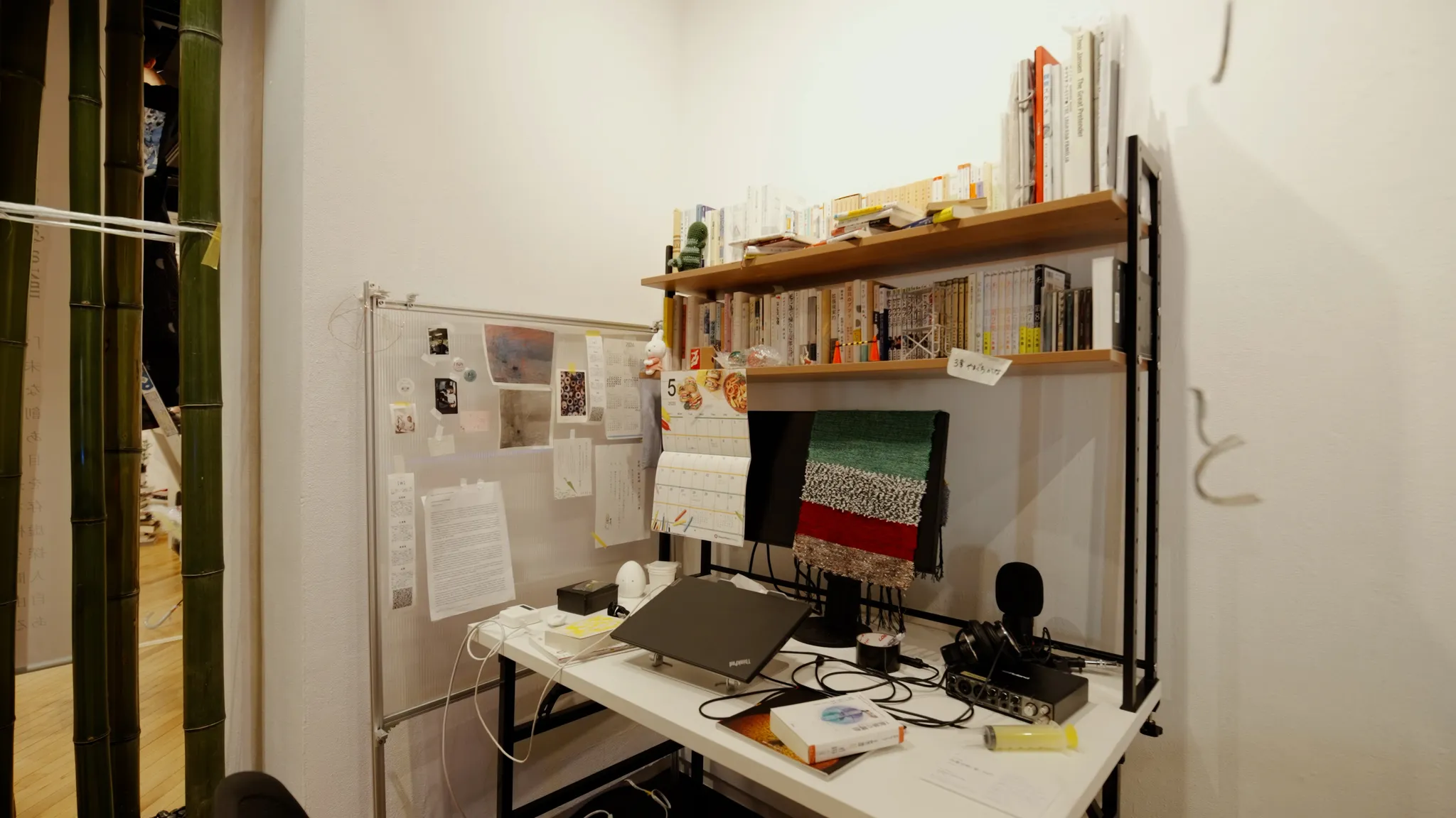 研究室のデスクを模した展示。デスク上にノートPC、モニター、カレンダーなどが立てられた様子。背後の壁には書類や本棚がある。