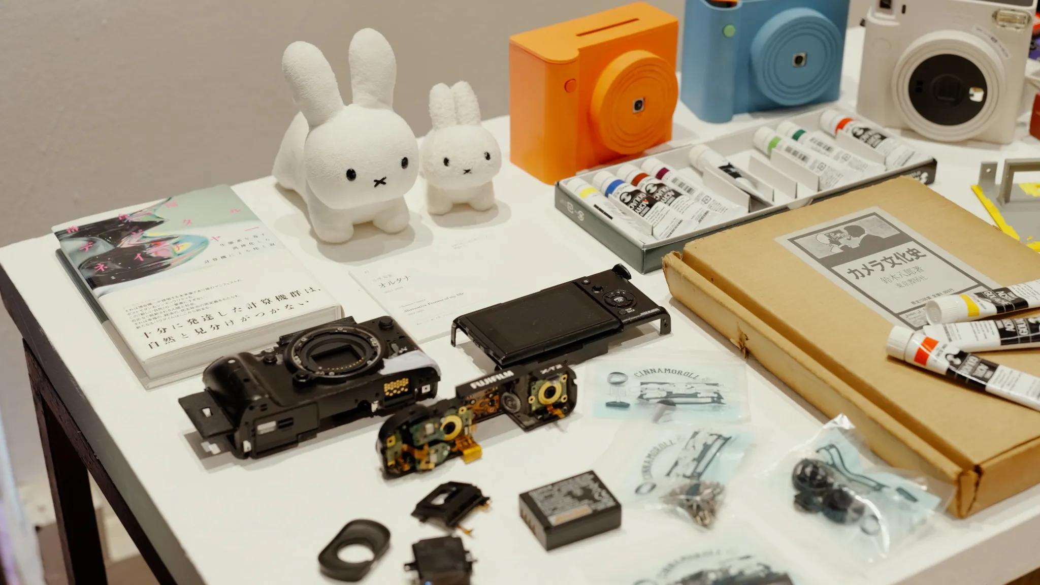 白いテーブルの上に、白いウサギのぬいぐるみ、オレンジと青のカメラ、カメラの分解パーツ、アクリル絵の具、本、茶色の箱が並んでいる。