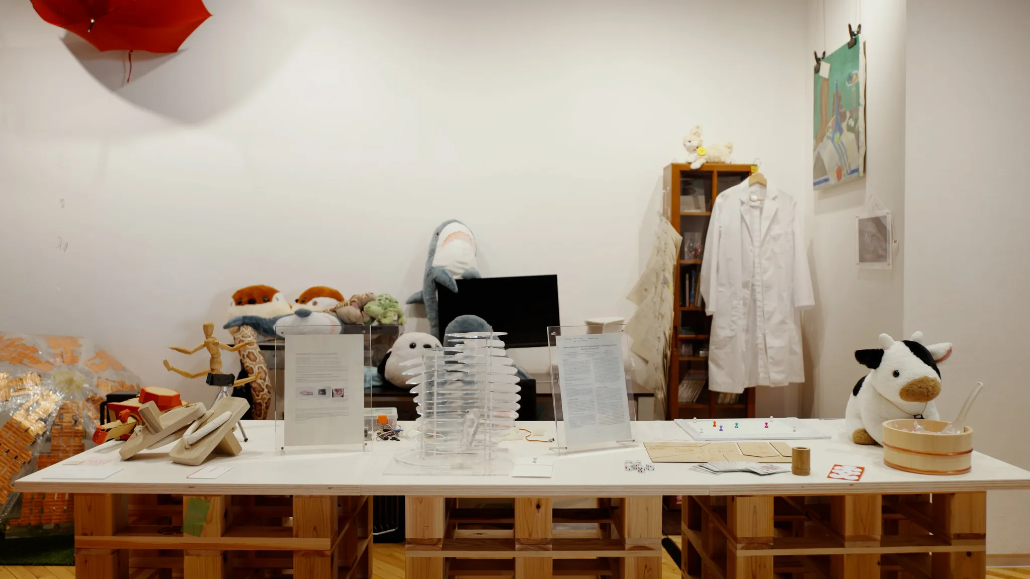 パレットで作られたテーブルの上に、サメや牛のぬいぐるみ、アクリルの展示物、書類、木製ゲタが並び、背後には白衣が掛かっている展示スペース。