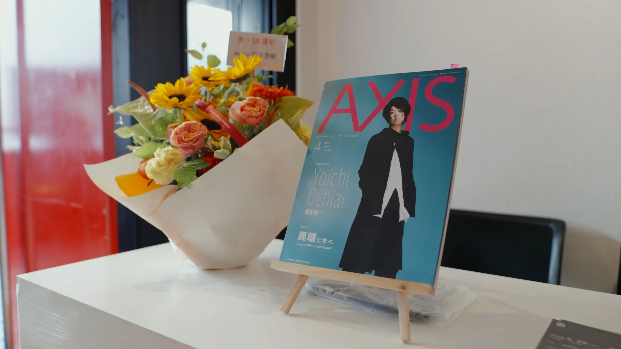 白い台の上に、ヒマワリの花束が入った白い花瓶と、落合陽一氏が表紙の雑誌「AXIS」が置かれている。