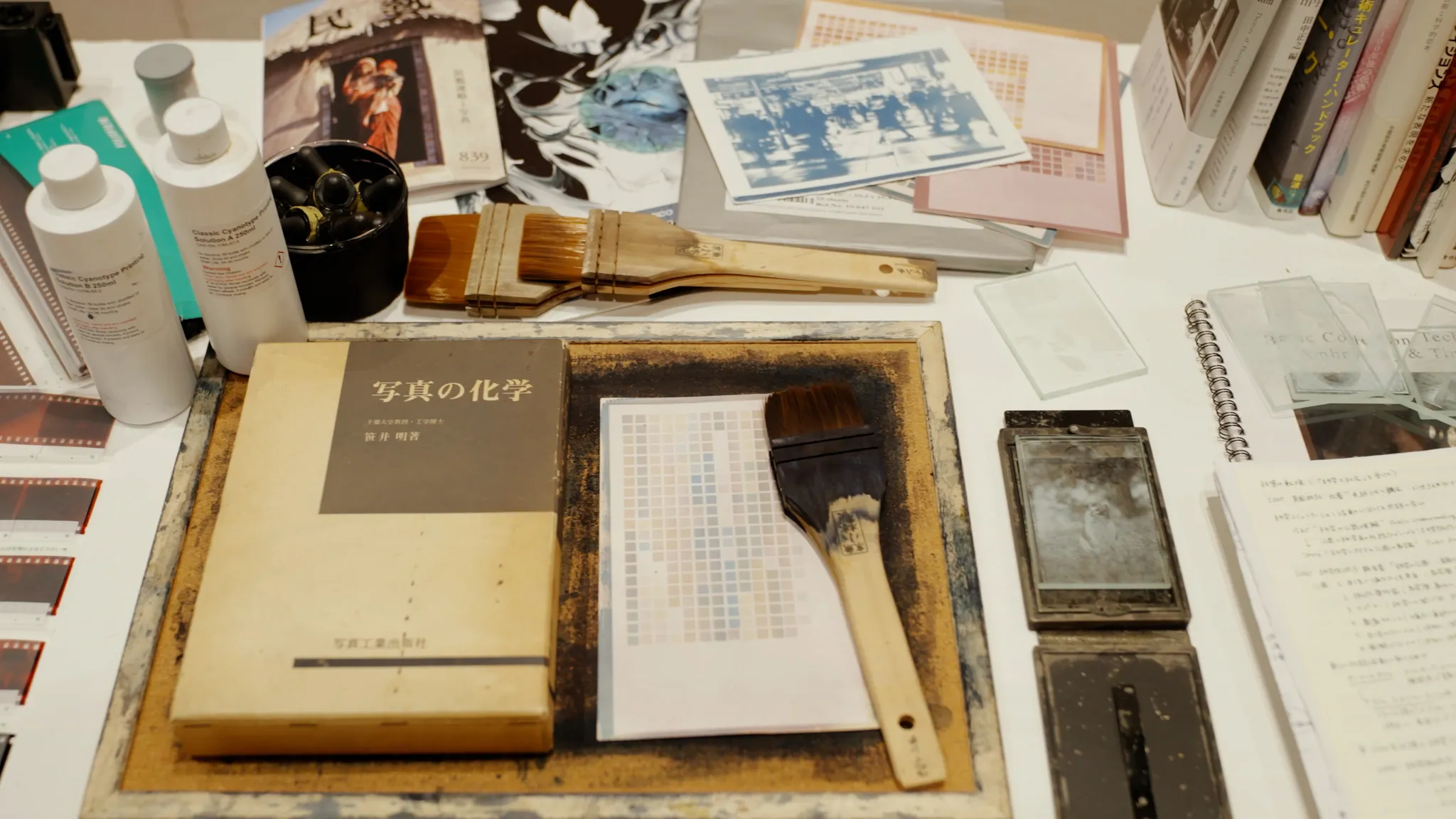 サイアノタイプの薬品ボトル、ハケ、「写真の化学」というタイトルの本、カラーチャートなど、写真の現像や制作に関わる様々な道具が机の上に広げられている様子。
