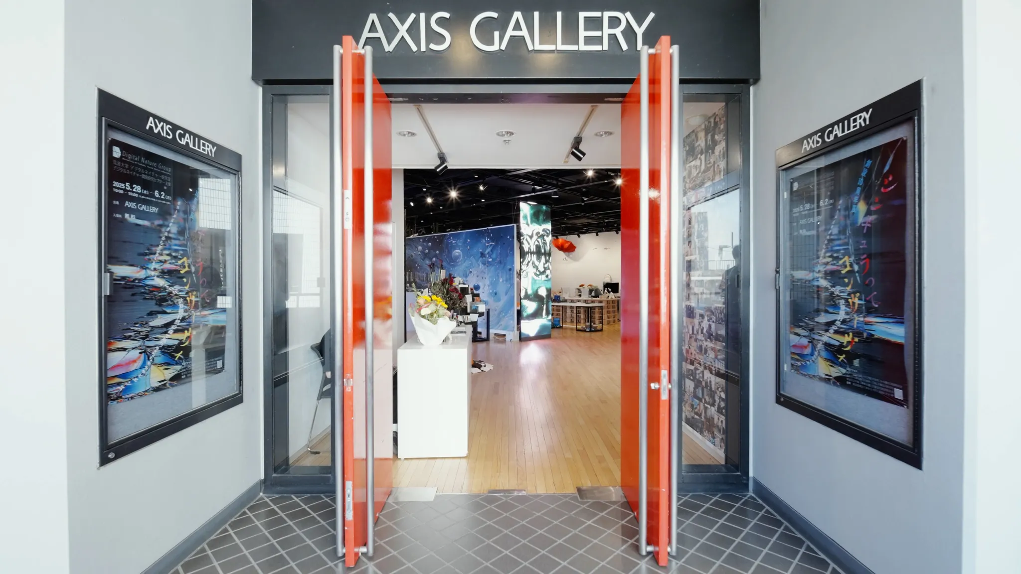 赤い扉が開かれた「AXIS GALLERY」のエントランス。入り口の両脇には展示会のポスターが掲示されており、奥にはギャラリー内の展示空間が広がっている様子が見える。