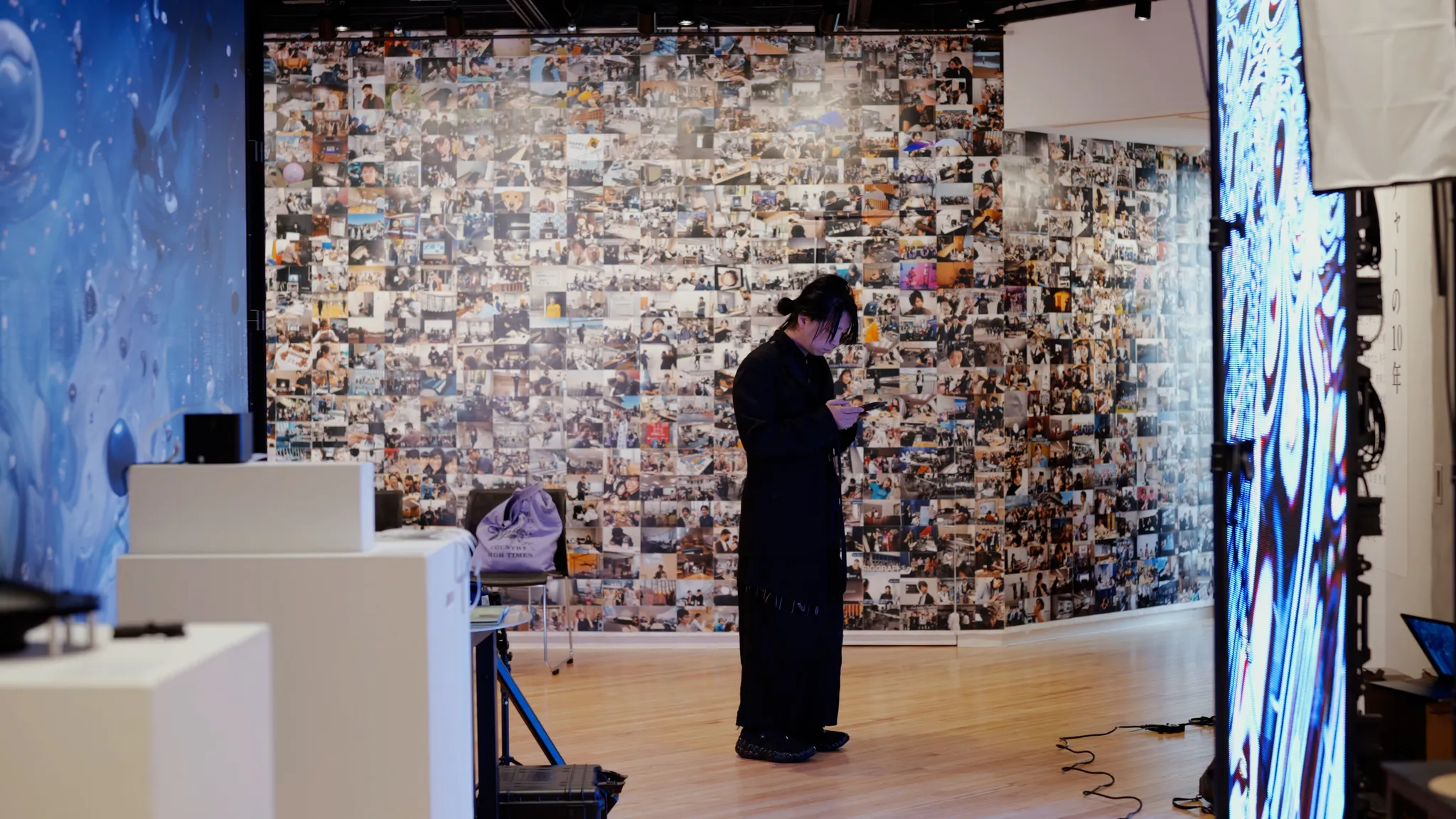 落合陽一氏が展示室でスマートフォンを操作している。背景には写真のコラージュ壁や、発光するデジタルディスプレイが見える。