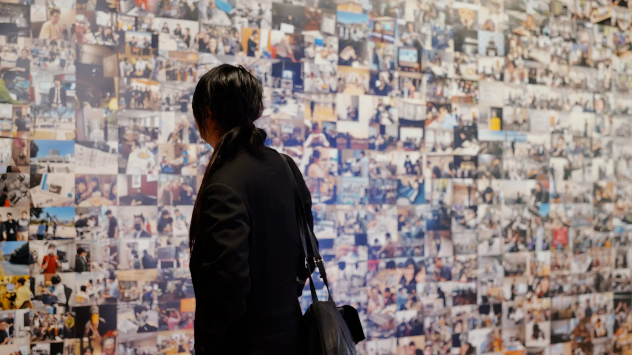 落合陽一氏が後ろ姿で立ち、小さな写真が隙間なく貼られた巨大なコラージュの壁を眺めている。