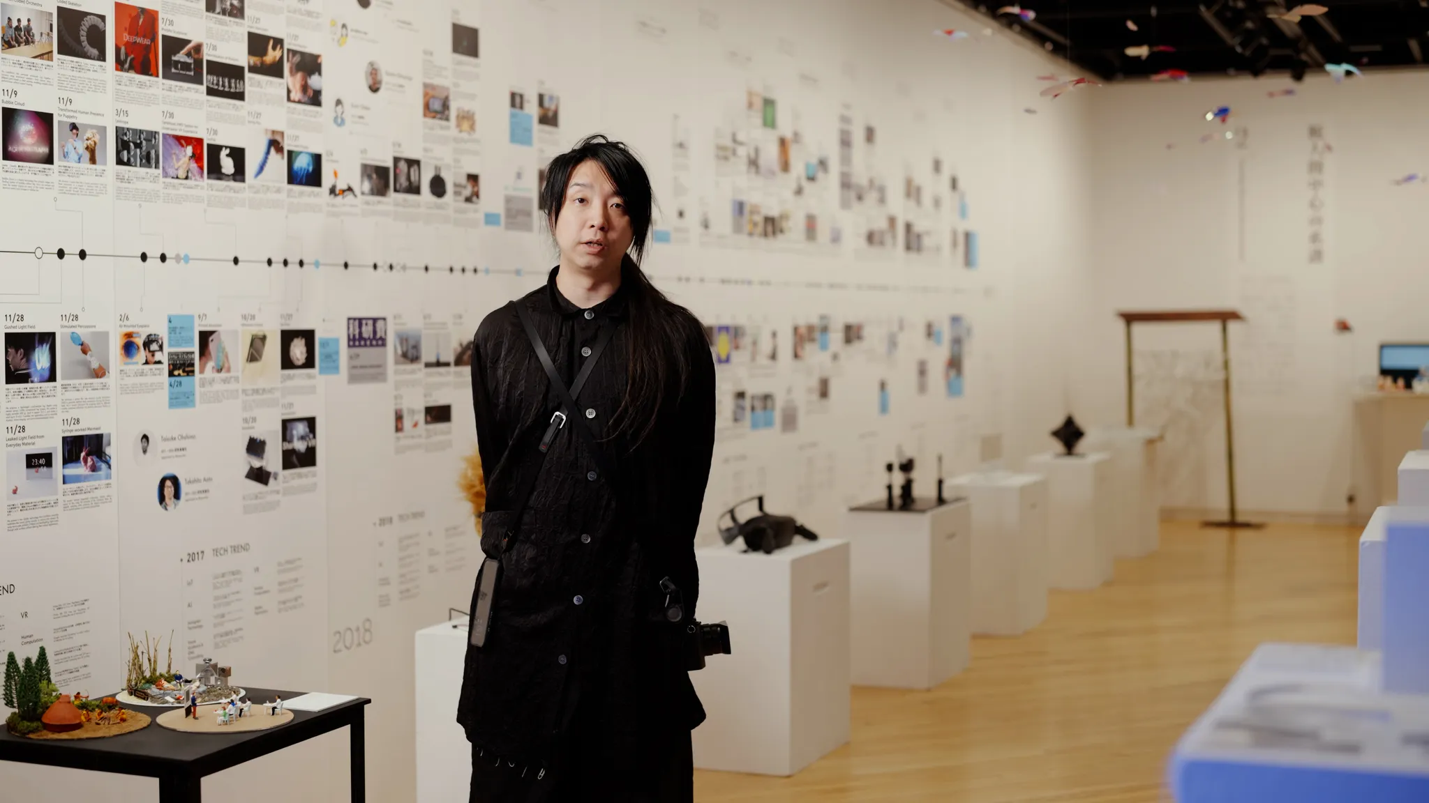 黒い服を着た落合陽一氏が展示空間に立っている。背景には、写真やテキストが配置された巨大な年表の壁と、展示物が置かれた白い台がある。