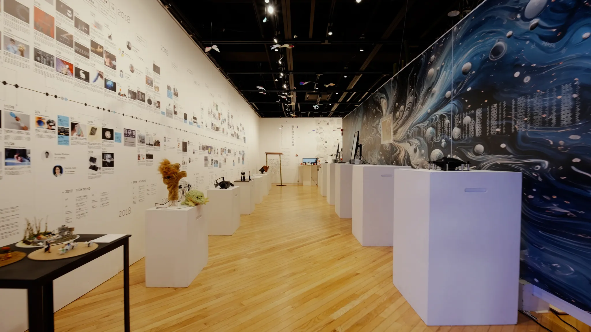板張りの床の展示空間の全景。左側の壁には長い年表があり、中央には展示物が置かれた白い台が並び、右側には抽象的なグラフィックのパネルがある。