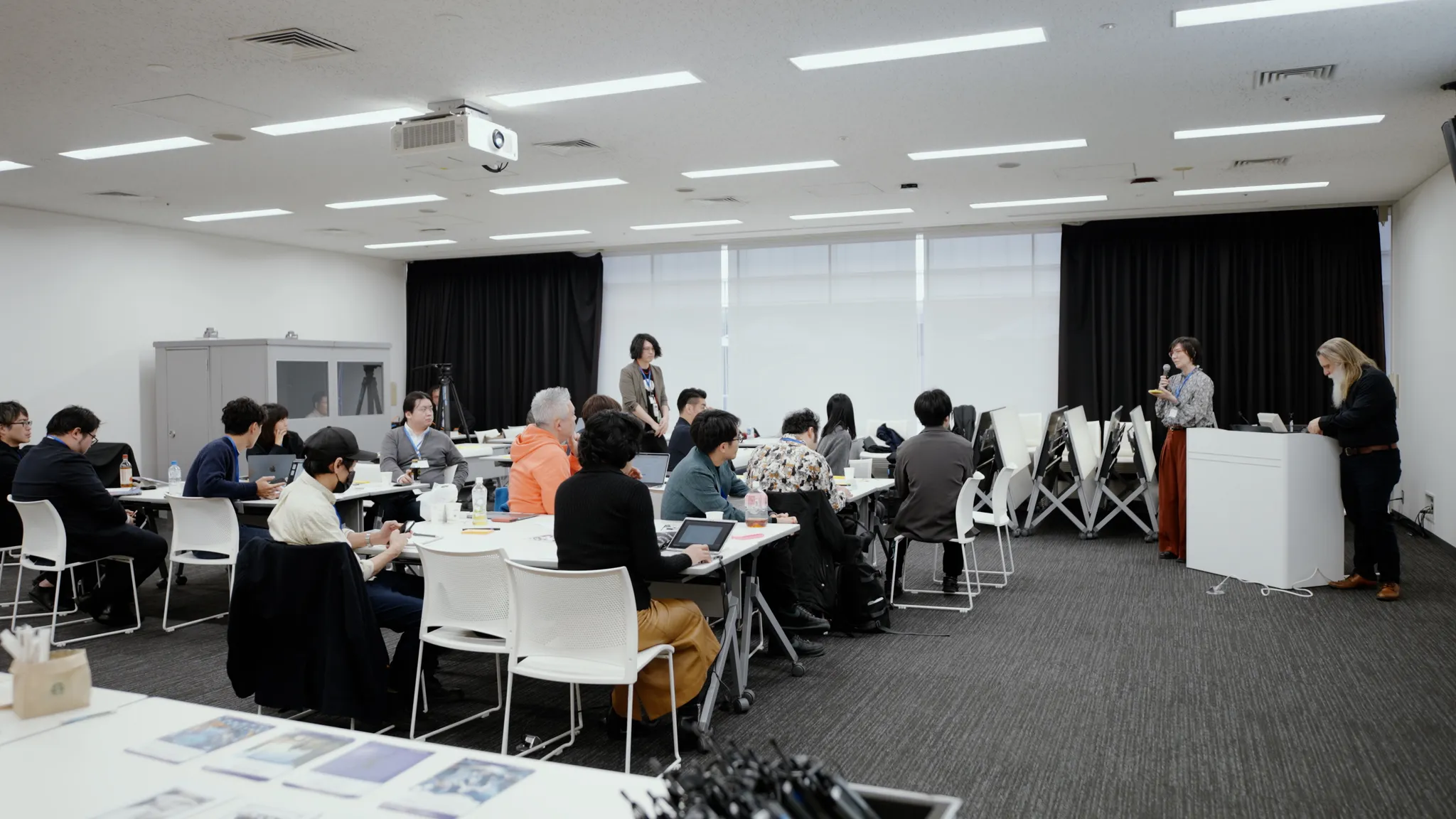 ワークショップの広いショット。参加者はテーブルに座り、演台に立つ発表者の方を向いている。