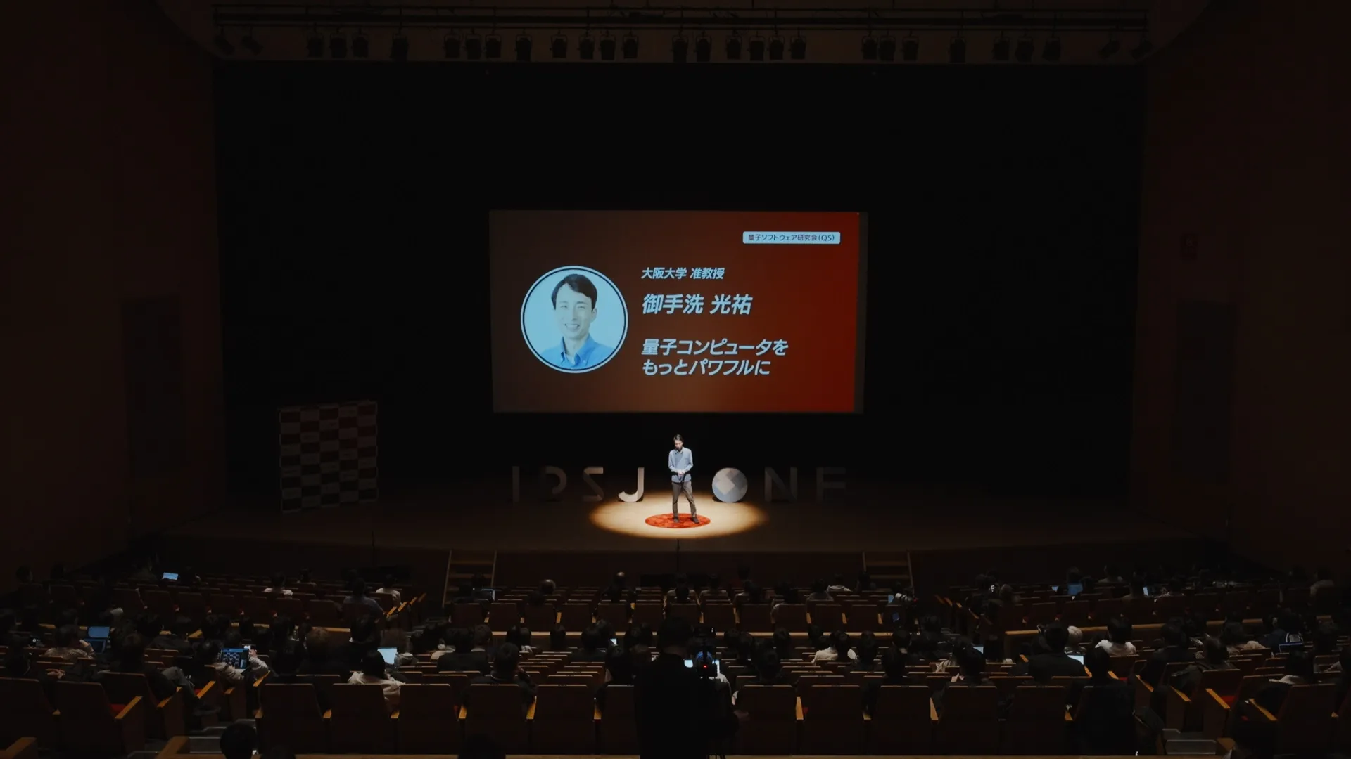 大きなホールで御手洗氏がステージ中央のスポットライトを浴びて立っている。背後のスクリーンには、登壇者の顔写真、名前「御手洗 光祐」、所属「大阪大学 准教授」、タイトル「量子コンピュータをもっとパワフルに」が表示されている。観客席は多くの人で埋まっている。