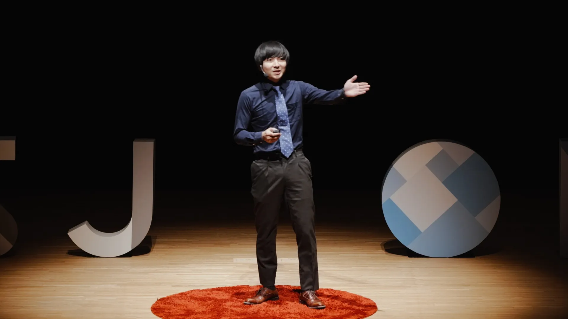 ヨビノリたくみ氏がステージ上でプレゼンテーションを行っている様子。黒いシャツと青いネクタイを着用し、左手を広げている。ステージには「IPSJ-ONE」のロゴの一部と円形のオブジェがある。