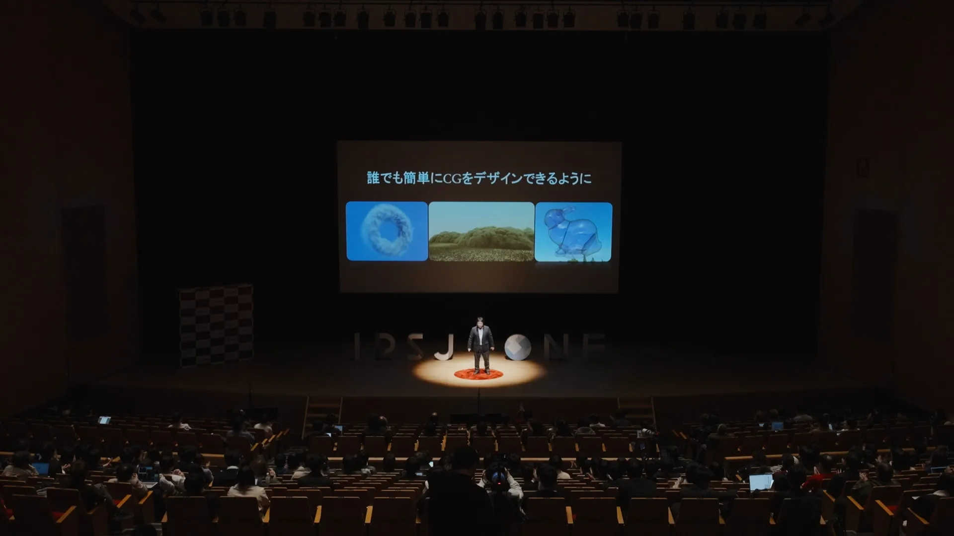 大きなホールで佐藤氏がステージ中央のスポットライトを浴びて立っている。背後のスクリーンには「誰でも簡単にCGをデザインできるように」というテキストと3つのCG画像が表示されている。観客席は多くの人で埋まっている。