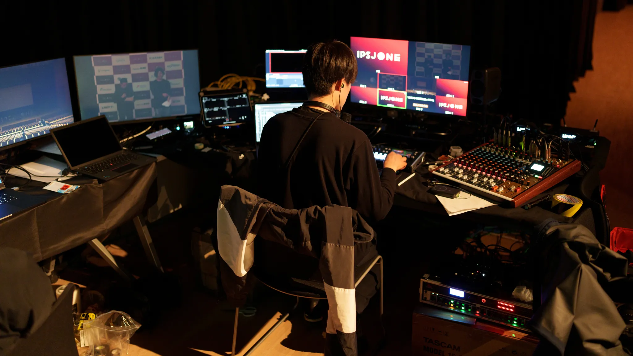 会場の技術スタッフが、複数のモニターとミキサーが並んだデスクで作業している後ろ姿。