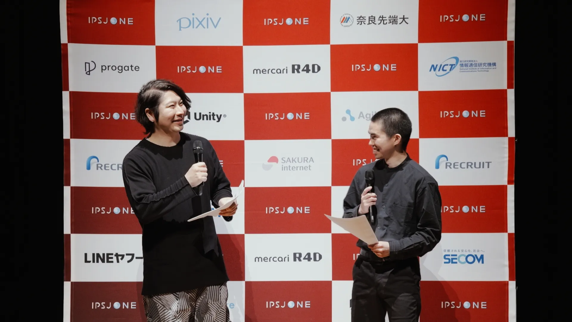 ステージ上でマイクを持って話している司会の2人。背景には「IPSJ-ONE」のロゴと多数のスポンサー企業のロゴが並んでいる。