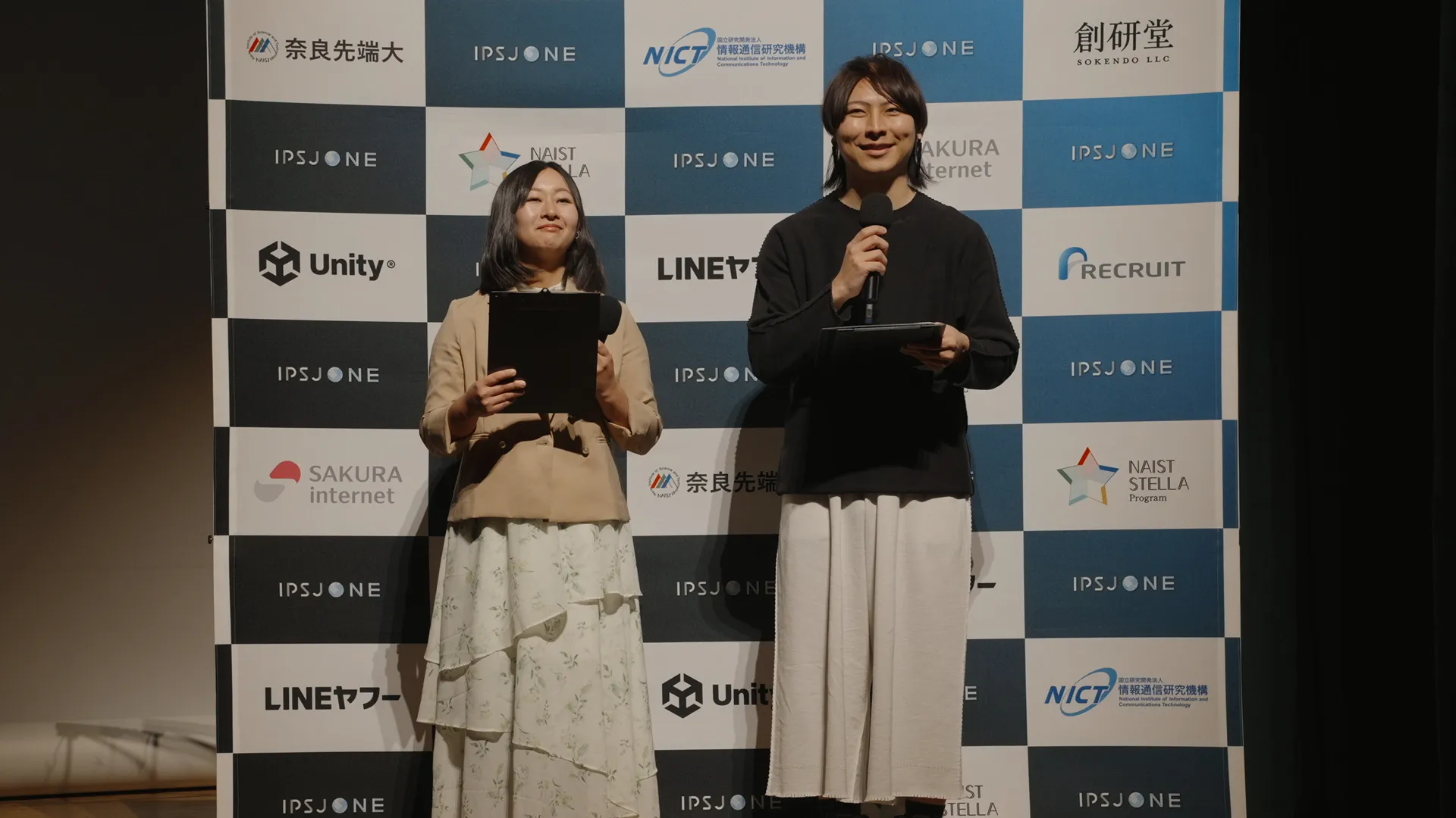 ステージ上でマイクを持って話している司会の2人。背景には「IPSJ-ONE」のロゴと多数のスポンサー企業のロゴが並んでいる。