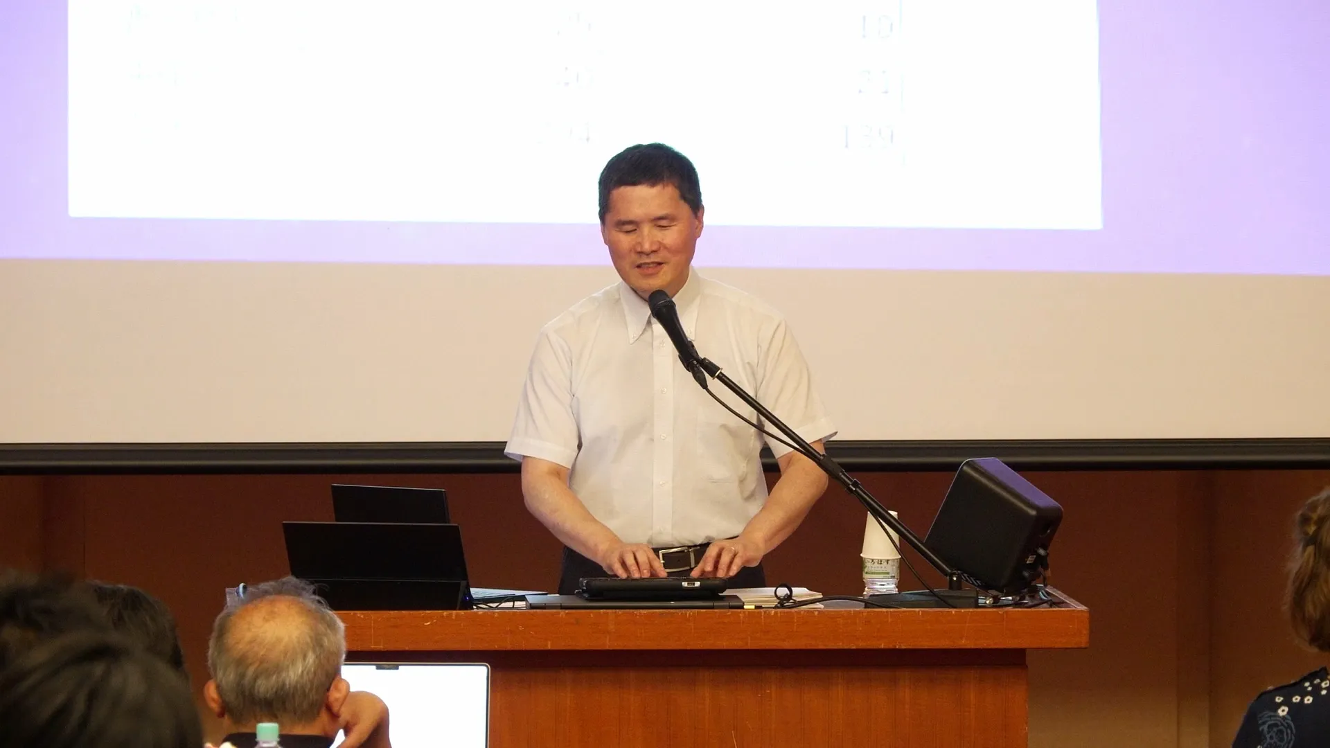 演台に立つ発表者がマイクに向かって話している。背後にはプロジェクタースクリーンが映っている。