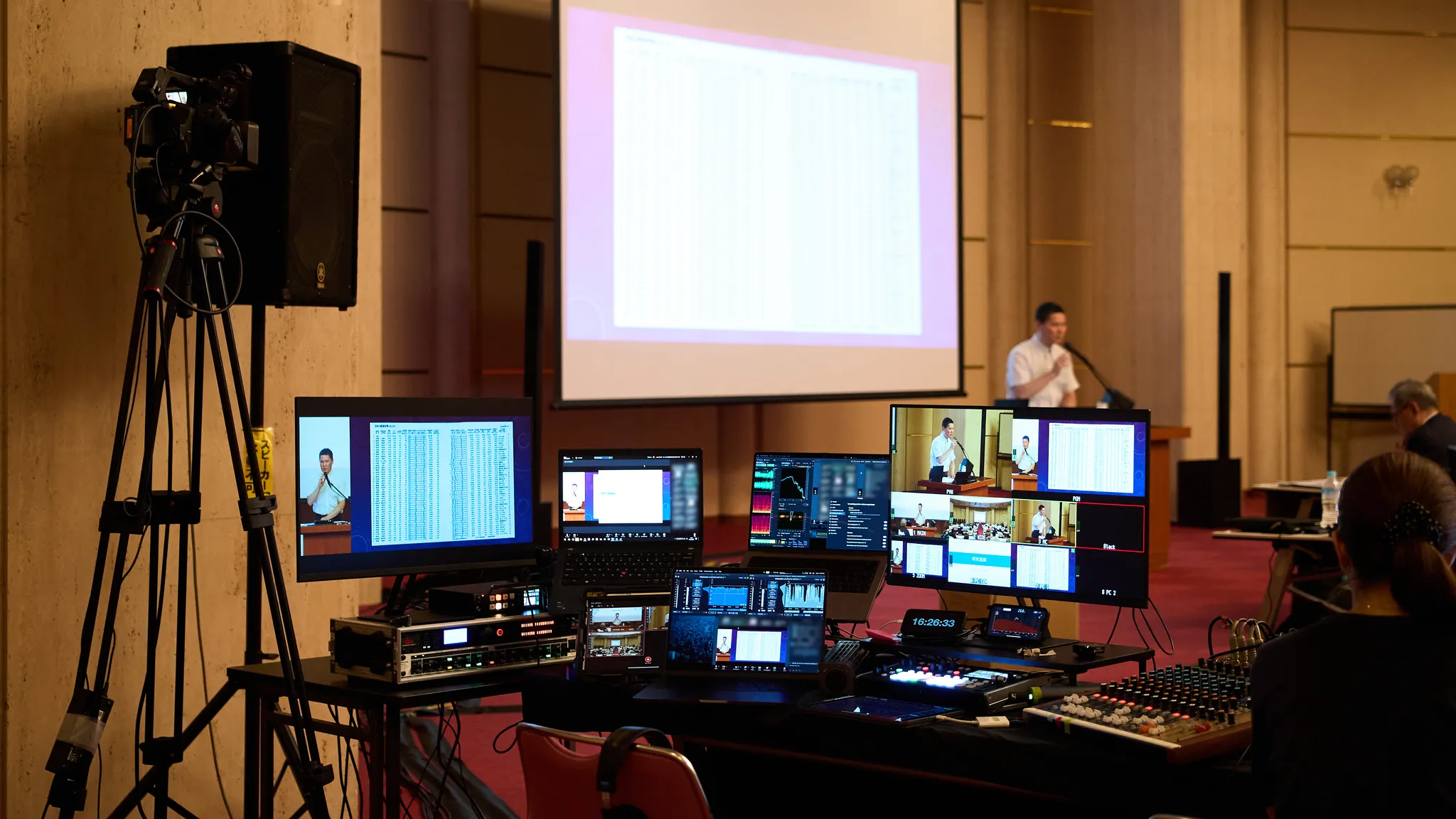 複数のモニターやミキサーが並ぶイベントの映像・音響ブース。奥に発表者とスクリーンが見える。
