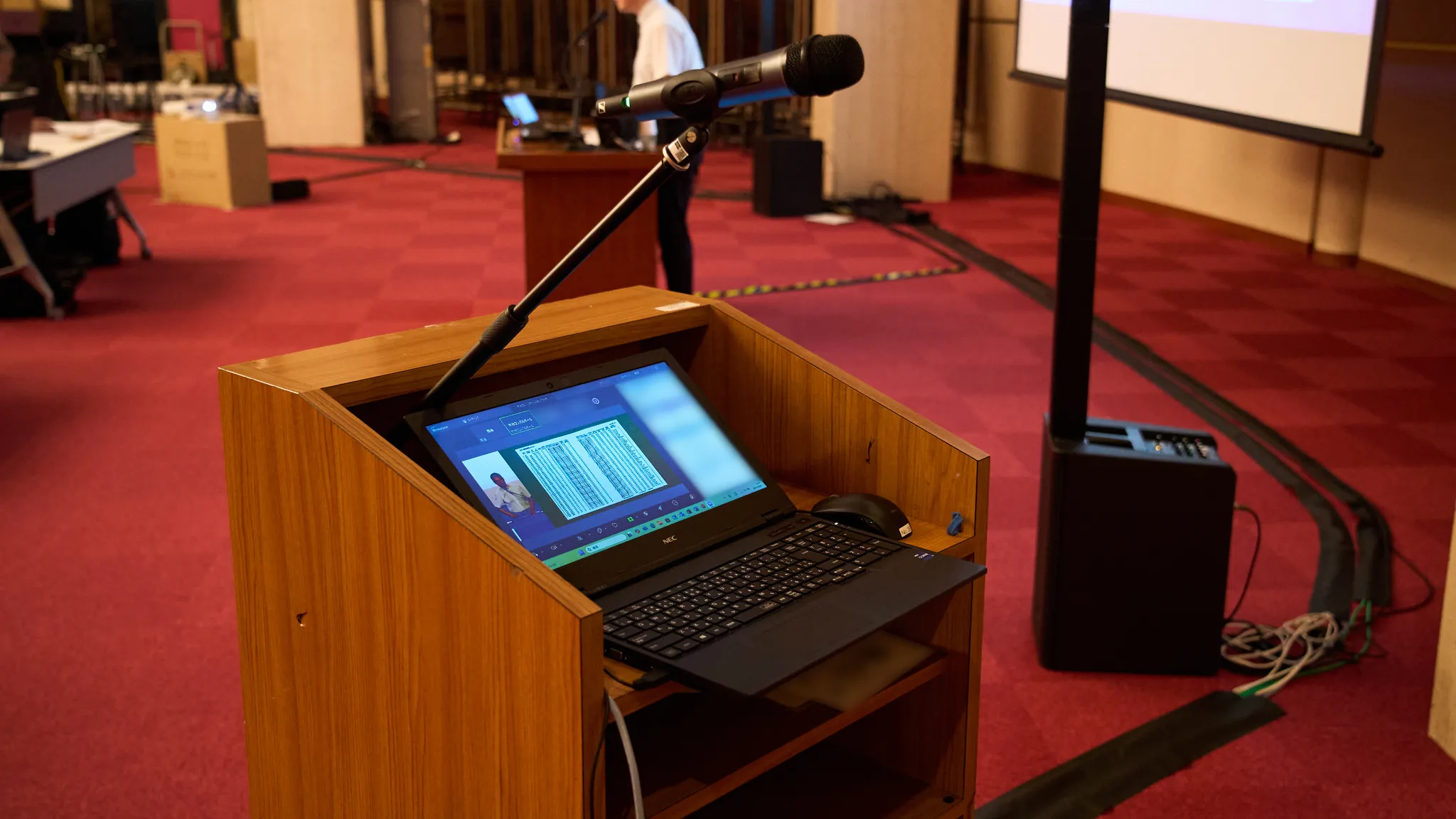 司会者用の演台に置かれたノートパソコンとマイク。ノートパソコンの画面にはZOOMビデオ通話の様子が映っている。
