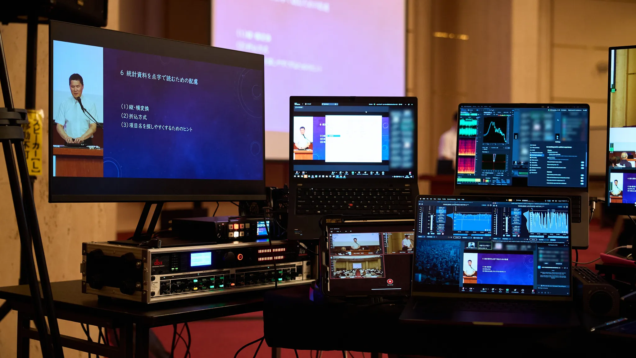 複数のモニターとラップトップが、発表者の映像やスライド、技術的な設定画面を表示している配信卓の映像・音響ブースの写真。