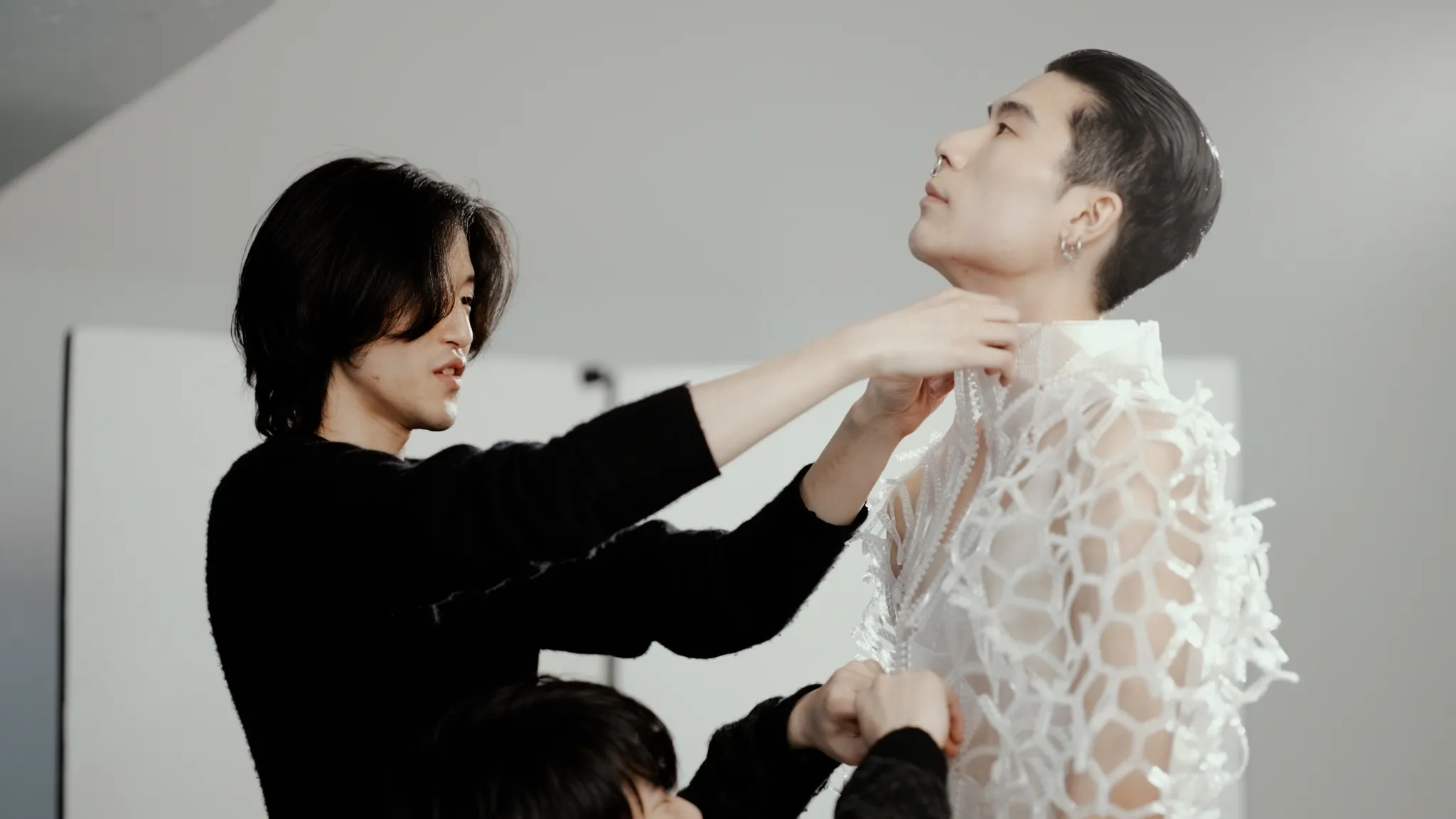 黒い服を着た人物（金田 昌也）が、白い網目状の複雑な衣装を着た別の人物の首元を調整している。