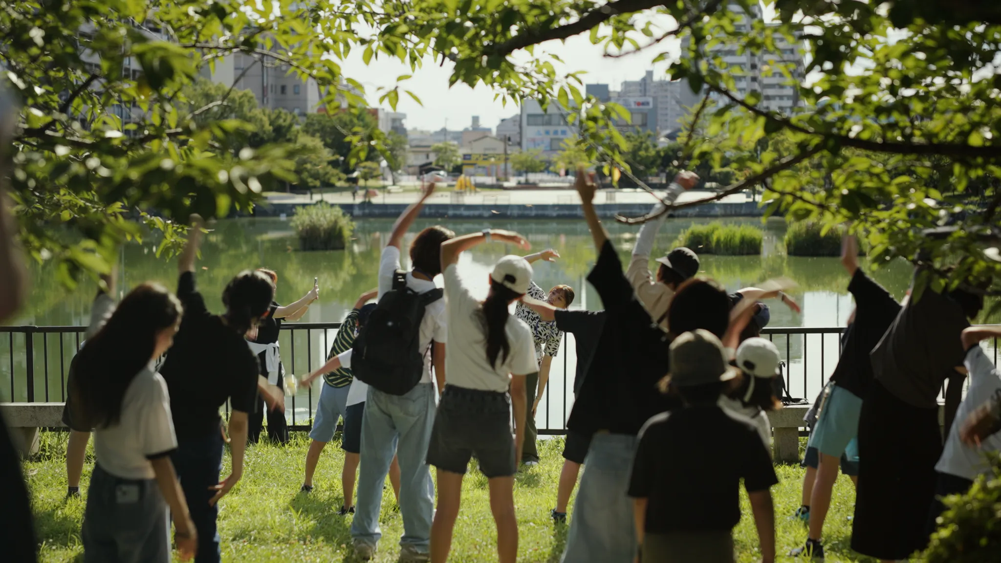 公園の木々の下で、参加者たちが水辺の芝生の上で腕を上げて体を動かしている。