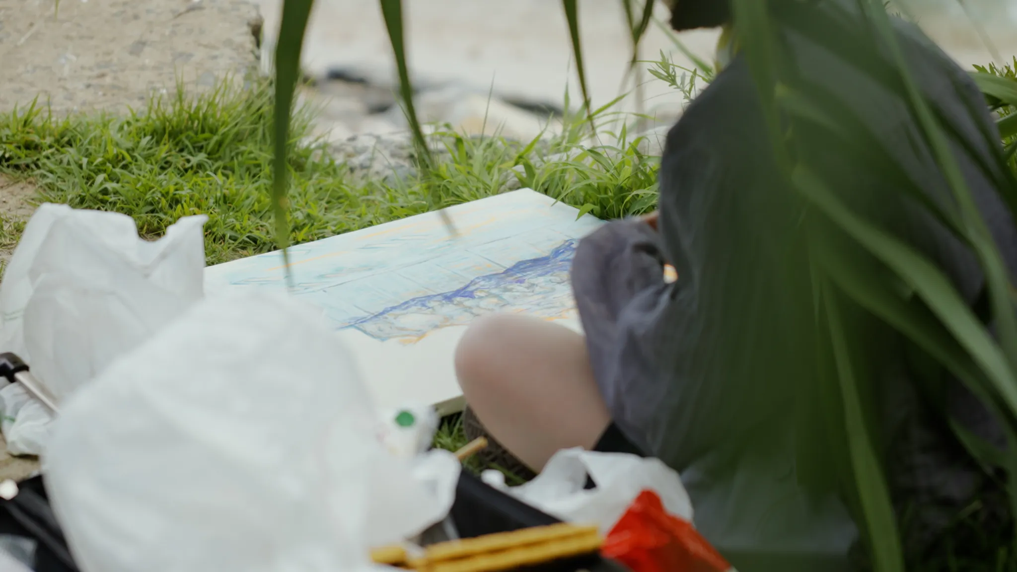 大きな葉に部分的に隠れながら、草の上に座ってキャンバスに絵を描いている人物。
