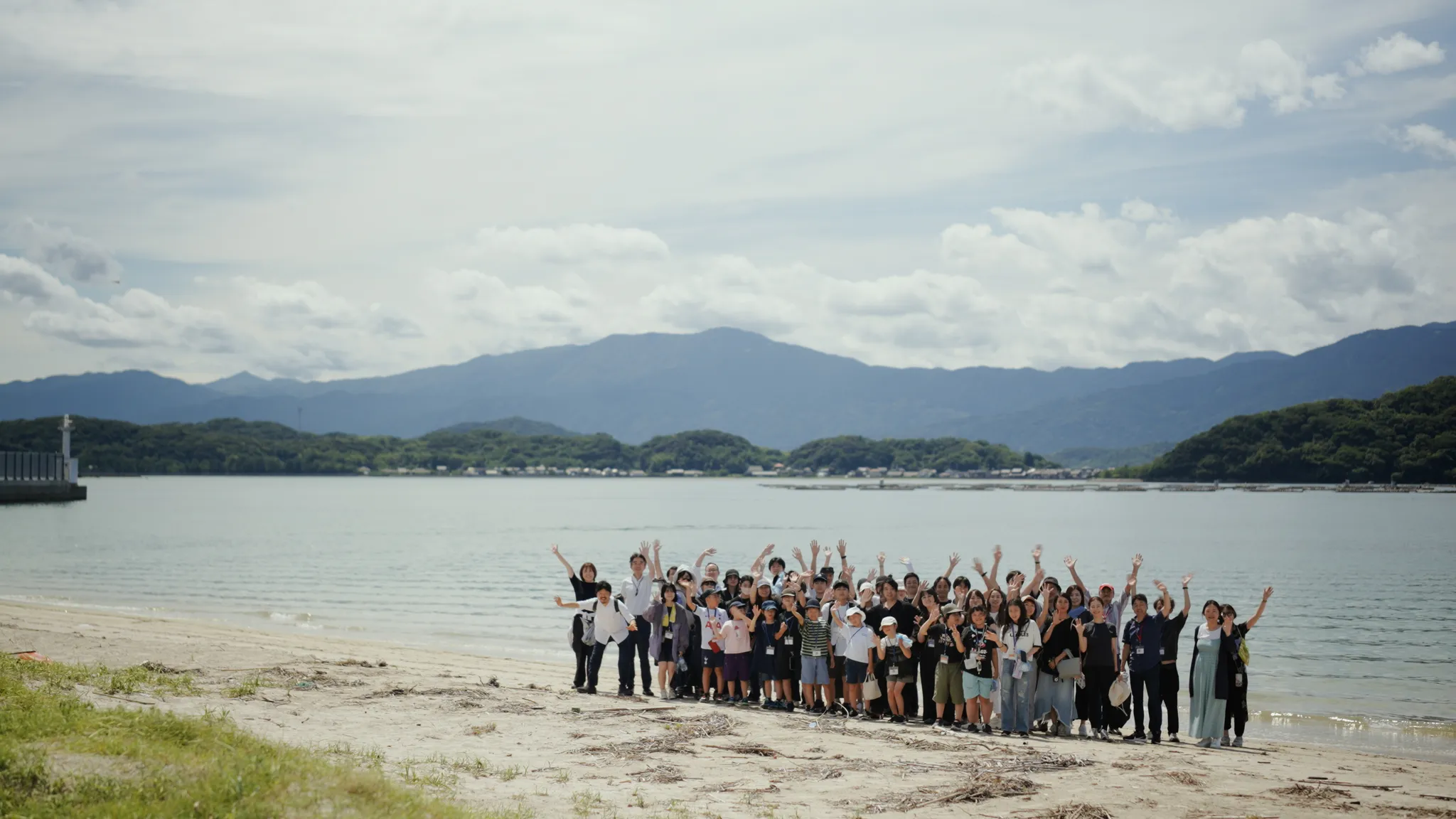 砂浜でポーズをとる人々の集合写真。後ろには海と山が広がっている。