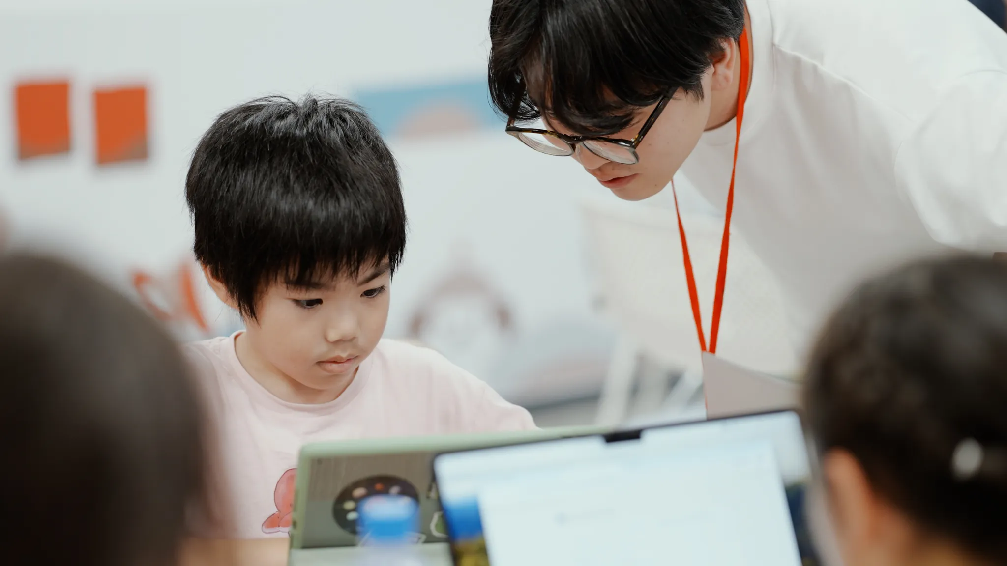 ティーチングアシスタントの大人がかがみ込み、タブレットを見ている子どもに何かを教えている。