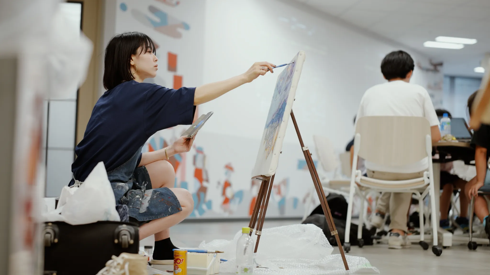 イーゼルに立てかけたキャンバスに、しゃがんで絵を描いているトノハルナ氏。