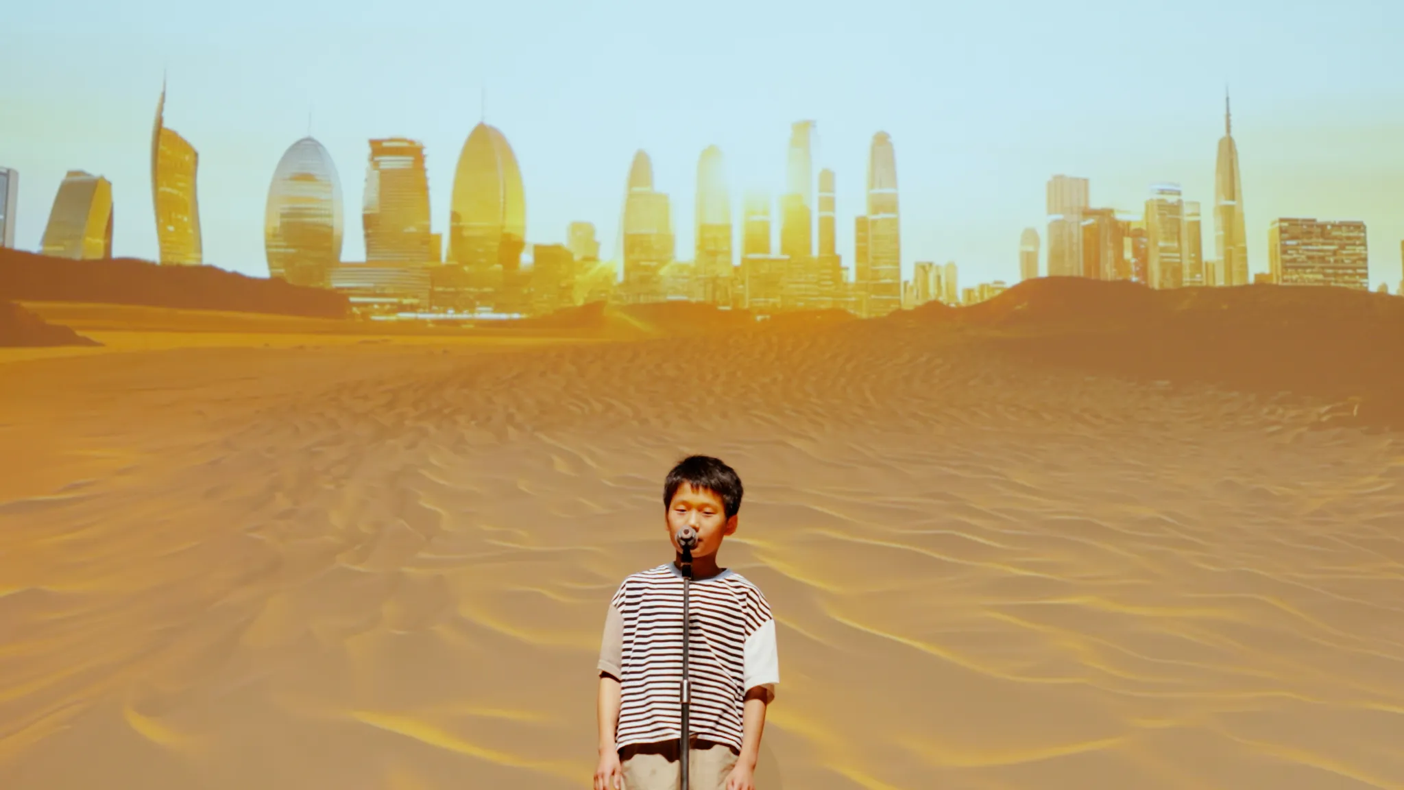 マイクの前に立つ子ども。背景には作品の砂漠と未来的な都市の風景が映し出されている。
