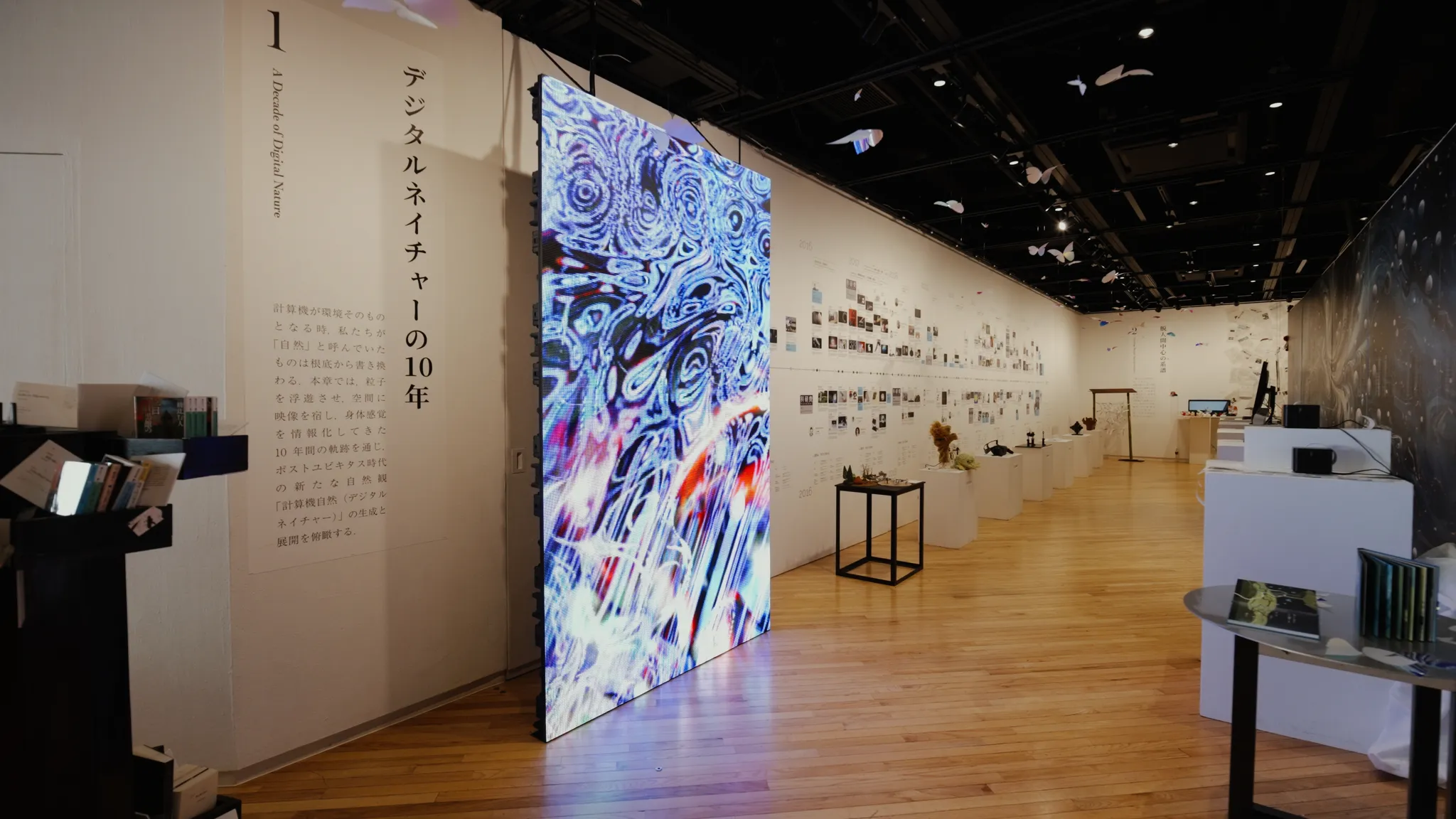 展示会会場の写真。「デジタルネイチャーの10年」と書かれたパネルがあり、中央にカラフルで抽象的なアートが映るLEDビジョン、奥の壁に年表と研究のアーカイブが並ぶ。