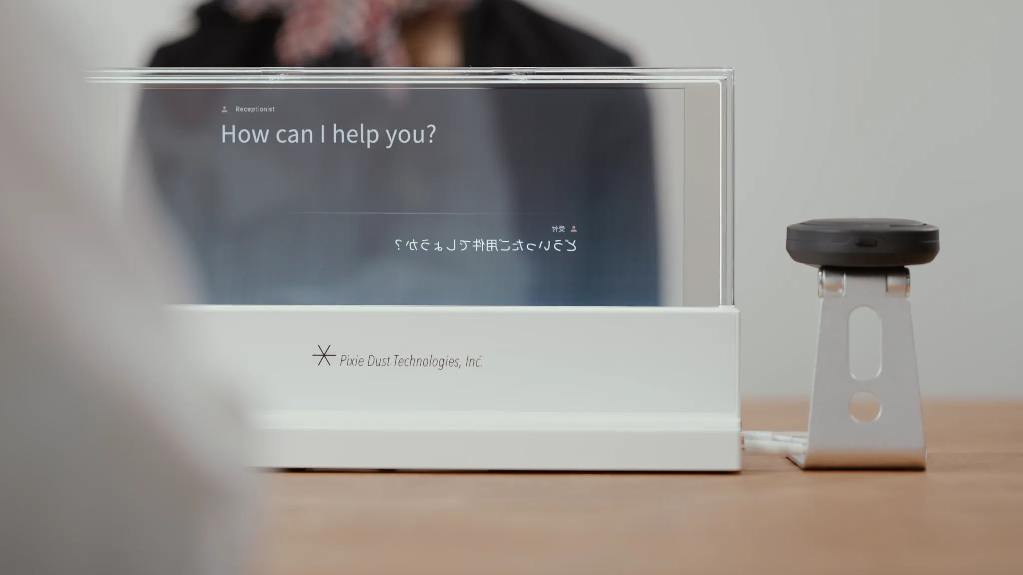 机の上に透明なディスプレイが置かれており、画面には「How can I help you?」という英語のテキストが表示されている。下部には日本語の反転文字で「どういったご要件でしょうか？」というテキストが表示されている。ディスプレイの下部には「Pixie Dust Technologies, Inc.」のロゴが記載されている。右側には小型のマイクがスタンドに設置されている。背景にはぼんやりと人物が映っている。