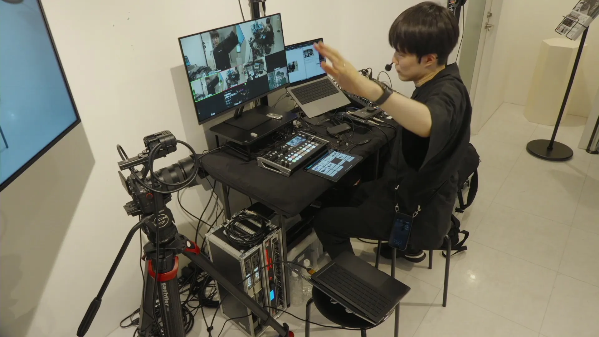 配信卓、複数のカメラ映像を表示するモニター、映像・音声機器を前に構成を解説する様子。