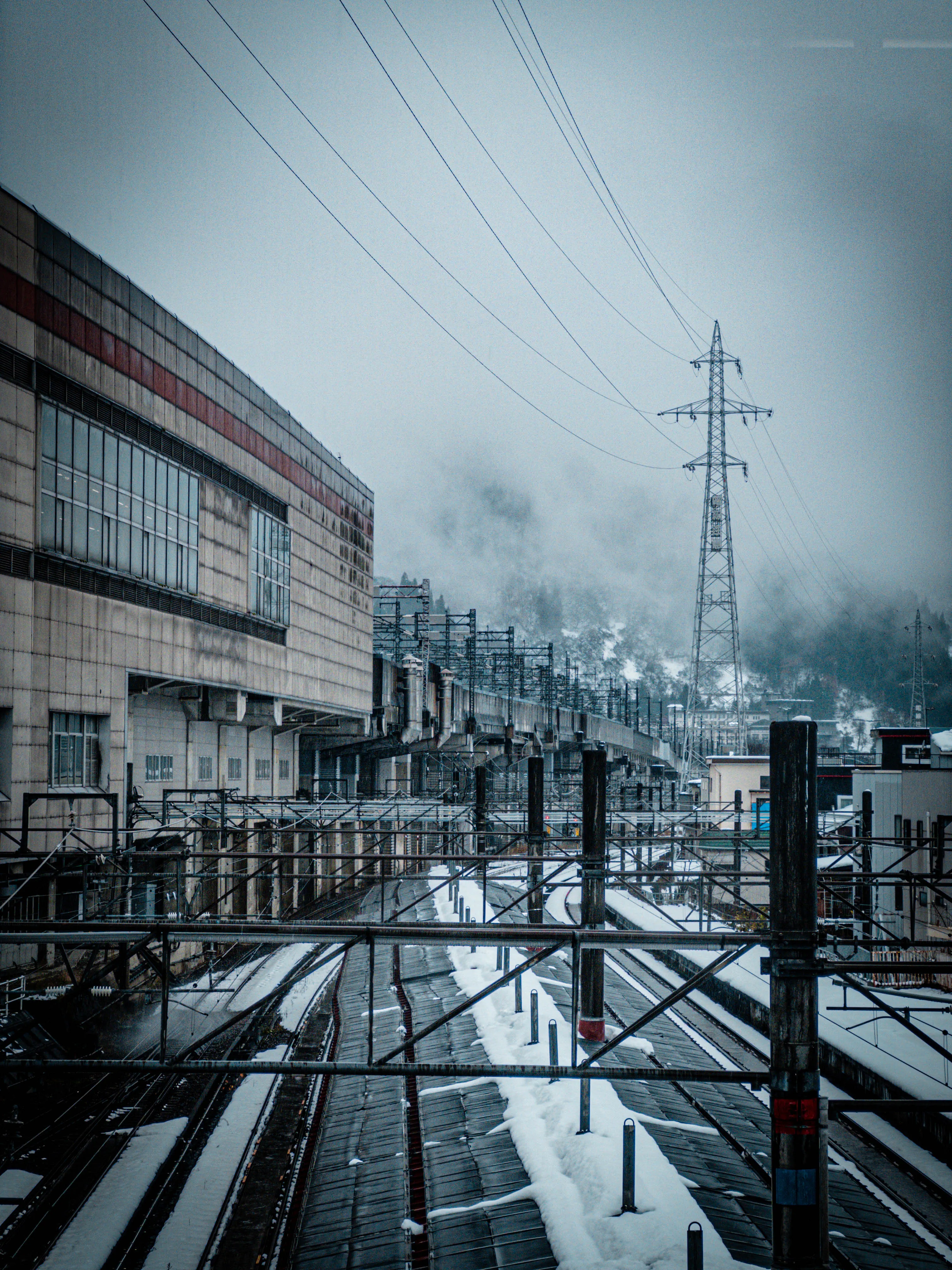 雪が積もった複数の線路と大きな建物、その奥には送電線の鉄塔が立ち込める霧と山を背景にそびえている冬の風景。