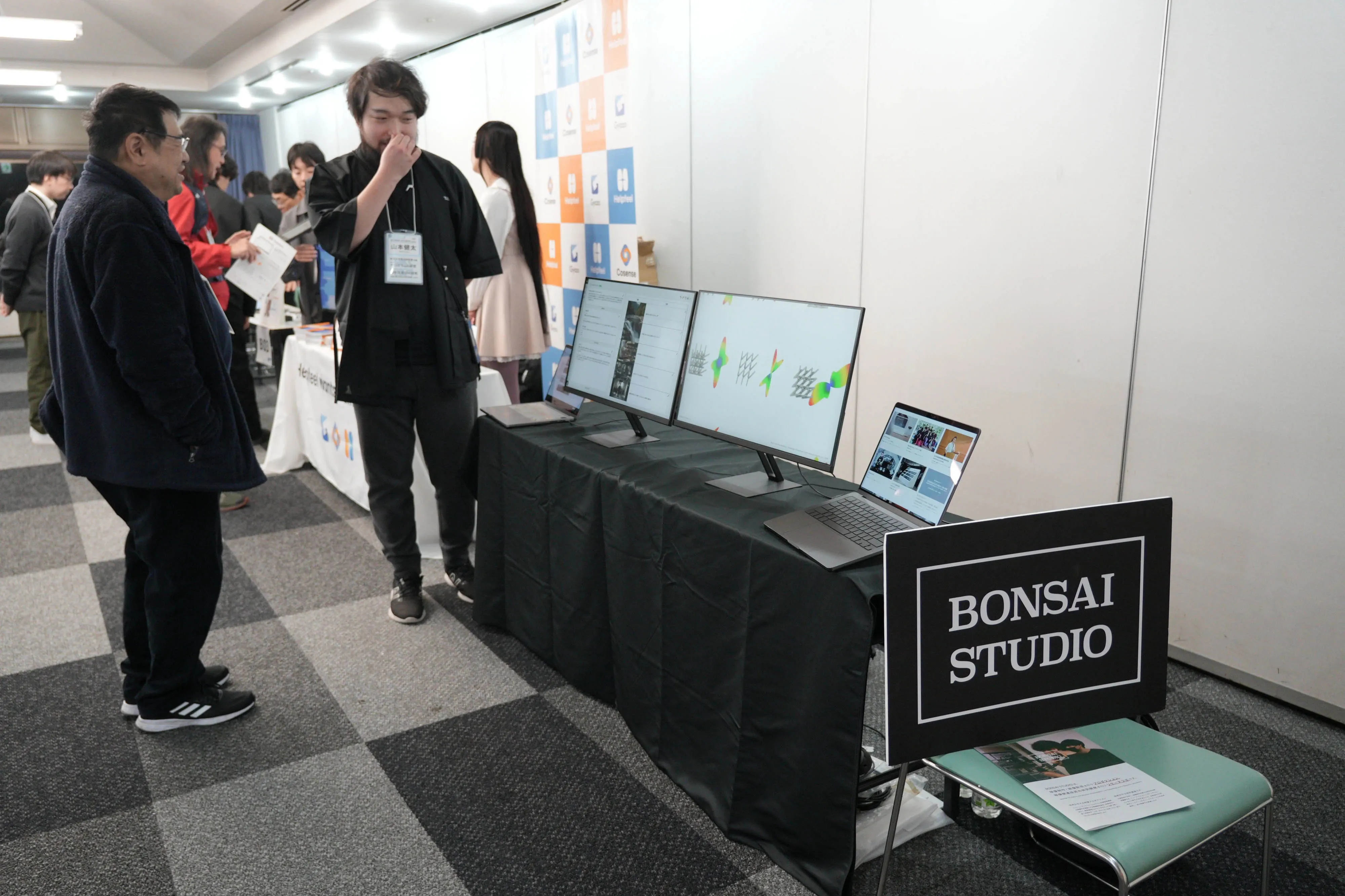 イベント会場の展示ブースでBONSAI STUDIOの看板を掲げ、黒い服を着たスタッフが複数のモニターを使って来場者に説明している。