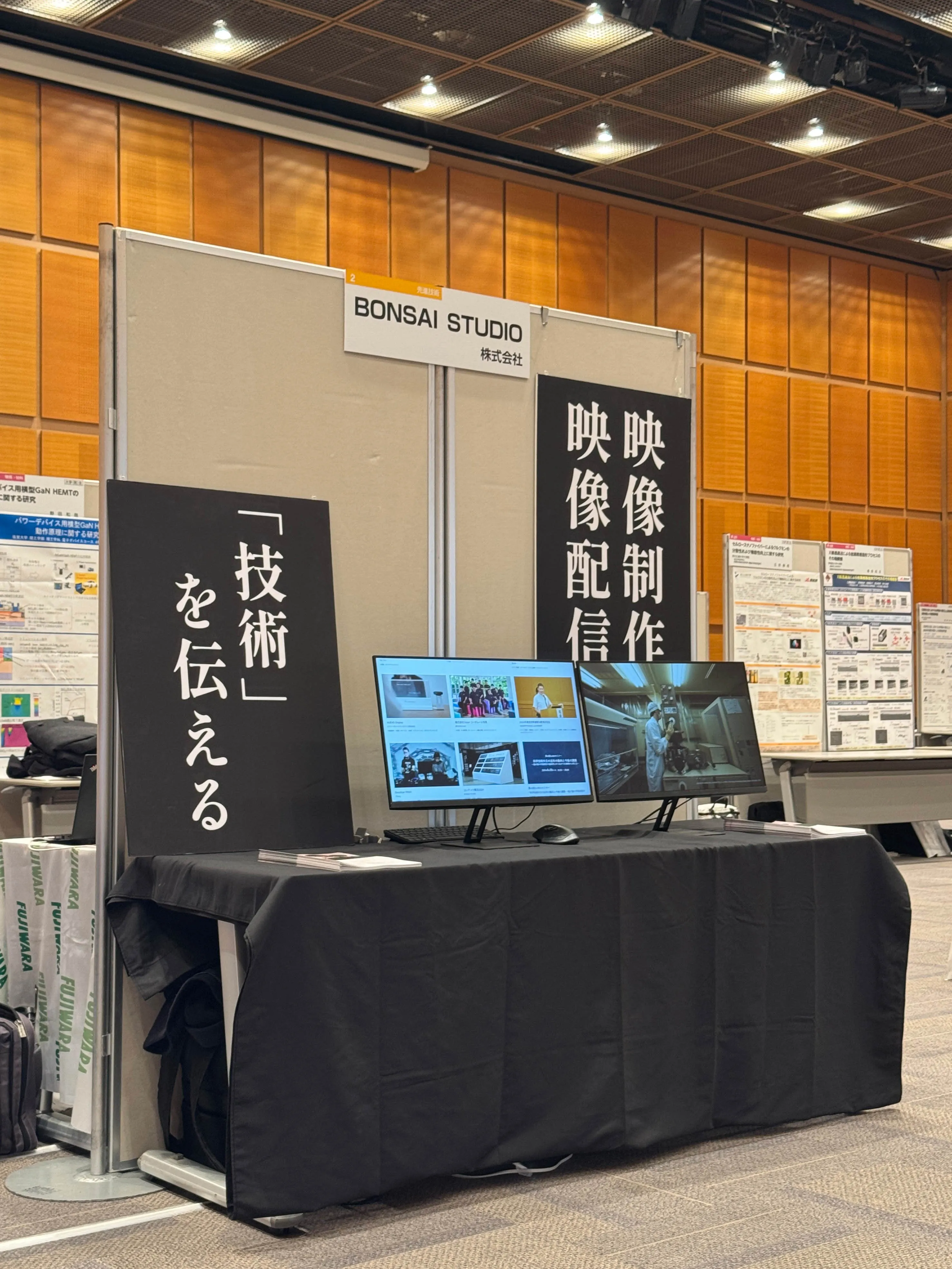 展示会場での「BONSAI STUDIO 株式会社」のブース。黒い布のかかったテーブル上にモニターが2台置かれ、大きな黒地のパネルには白字で「映像制作」「映像配信」「『技術』を伝える」などと書かれている。