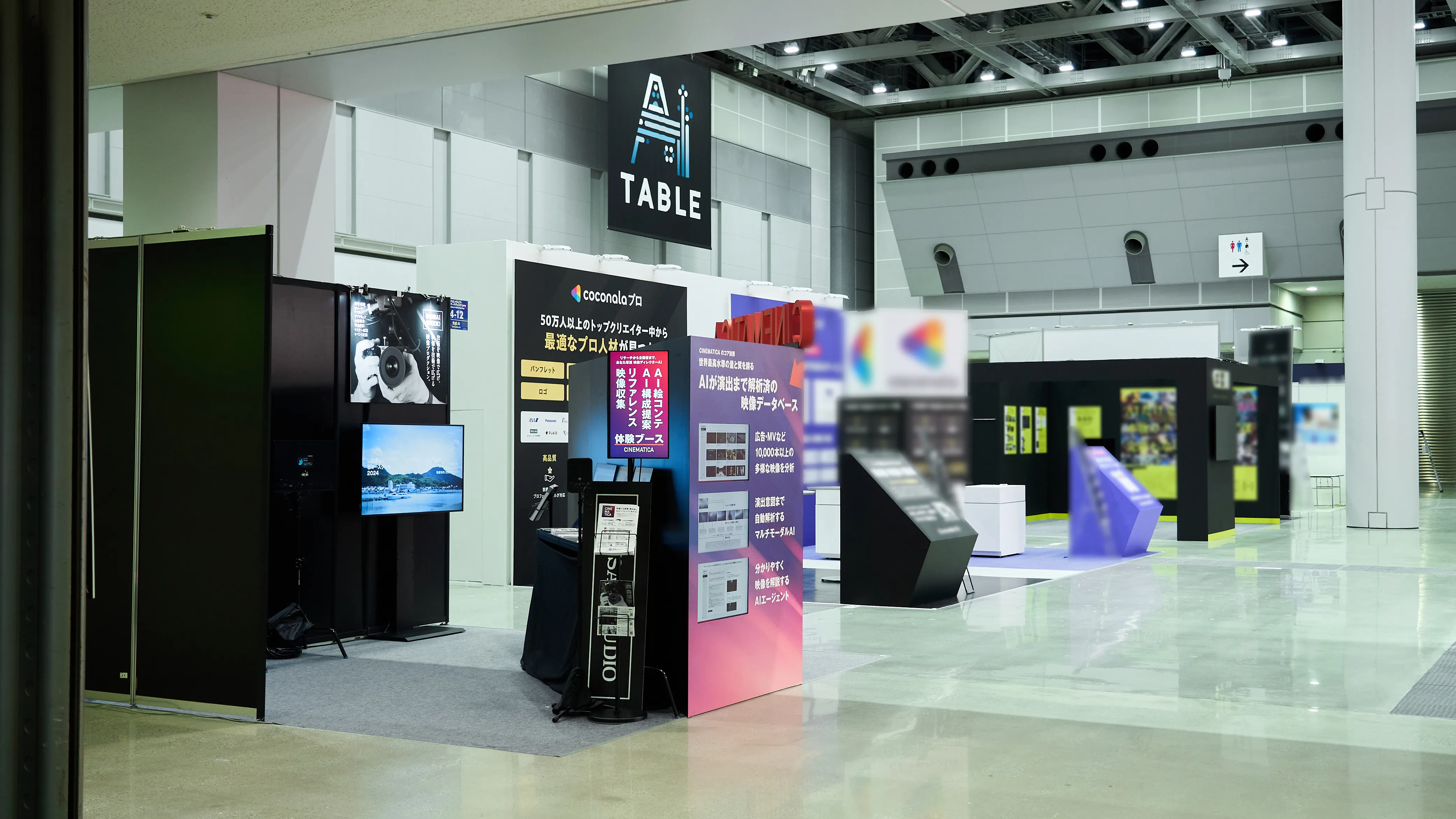 展示会場の通路からの風景。天井に「AI TABLE」のバナーが吊るされ、手前には「CINEMATICA」の赤いパネルのブース、奥には他社のブースが並んでいる。